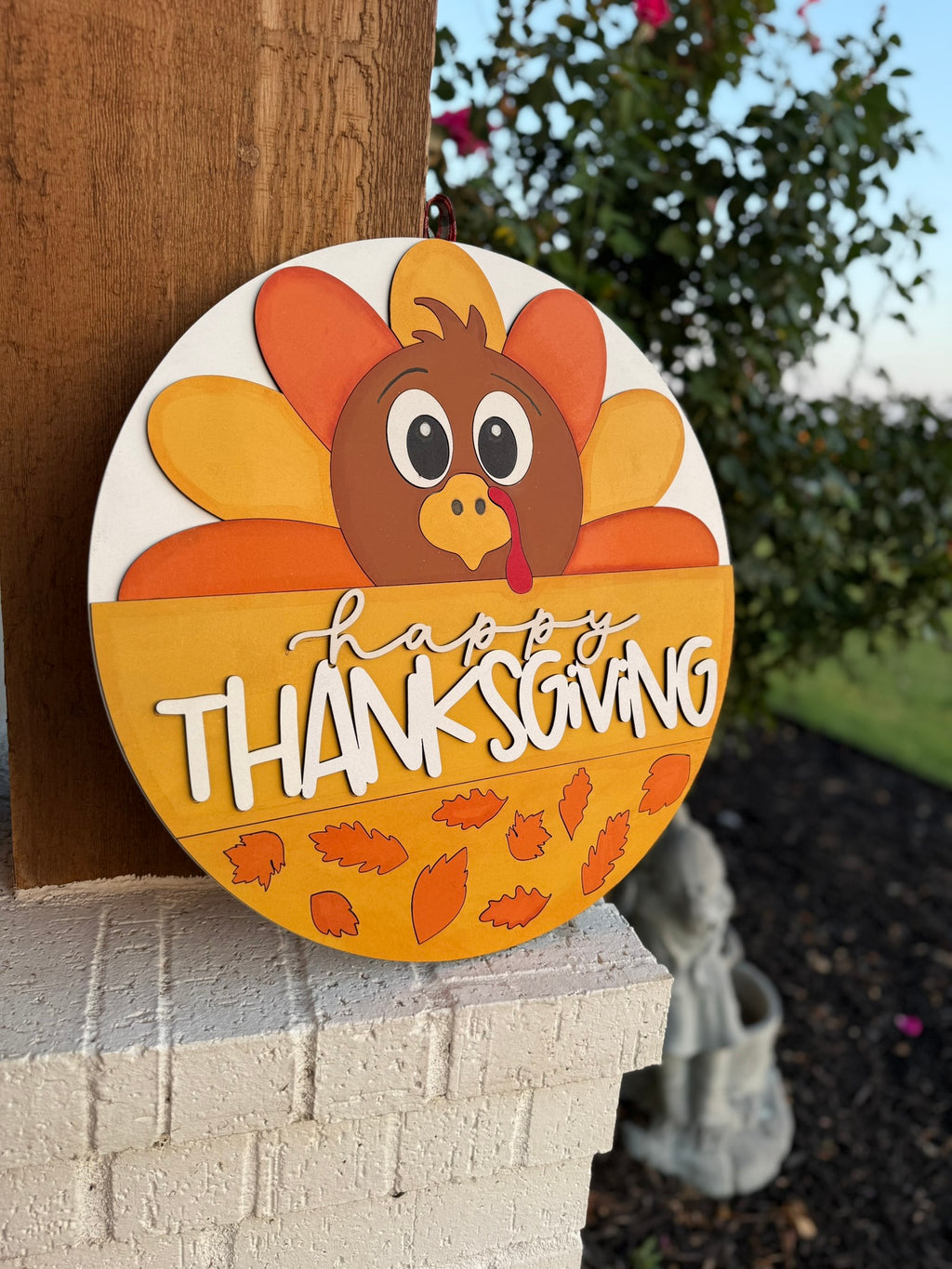 A round wooden sign with a cartoon turkey, colorful tail feathers, and the words Happy Thanksgiving, decorated with autumn leaves, hangs on a porch post with greenery in the background.