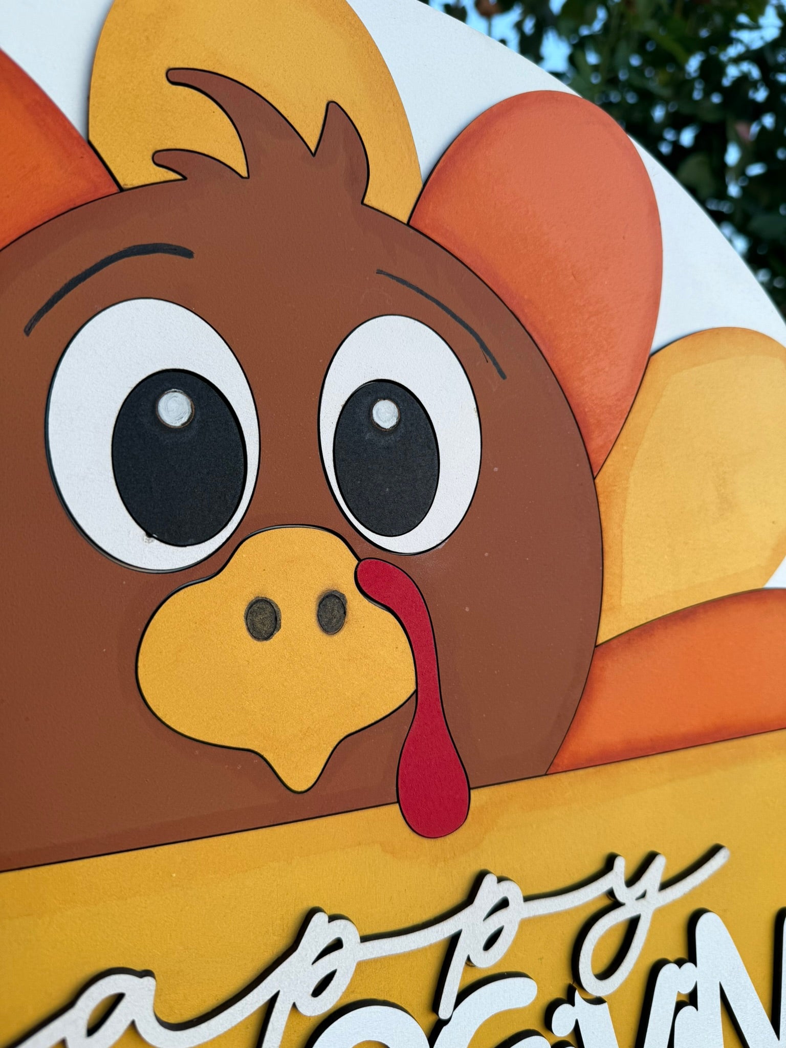 A close-up of a cartoon turkey face with large eyes, a yellow beak, and colorful feathers. The turkey has a red wattle, and part of the text Happy is visible at the bottom. Green leaves appear in the background.