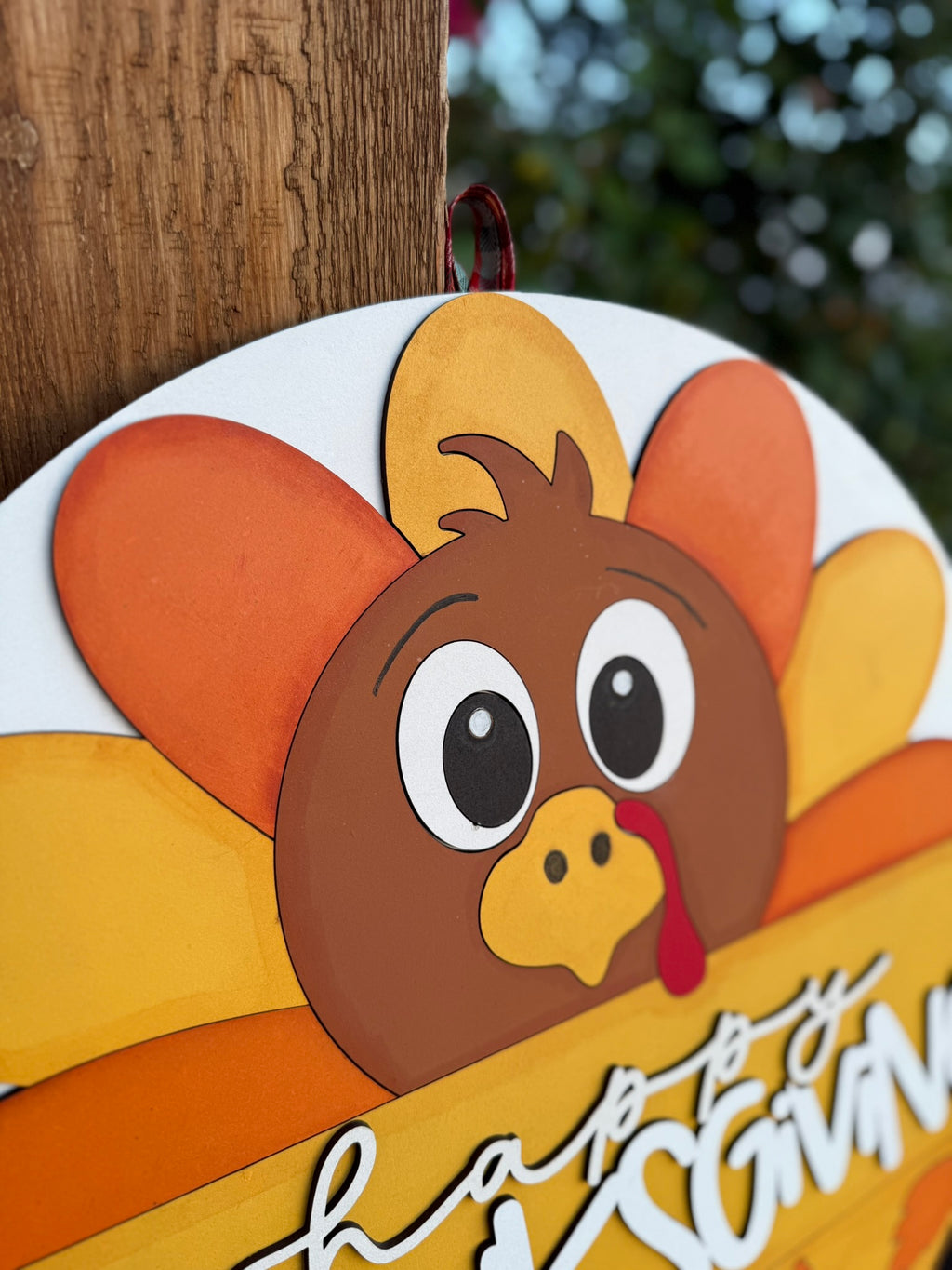 A close-up of a wooden Thanksgiving sign shaped like a cartoon turkey with colorful orange and yellow feathers, hanging on a post. The turkey has wide eyes and a red wattle. Greenery is blurred in the background.