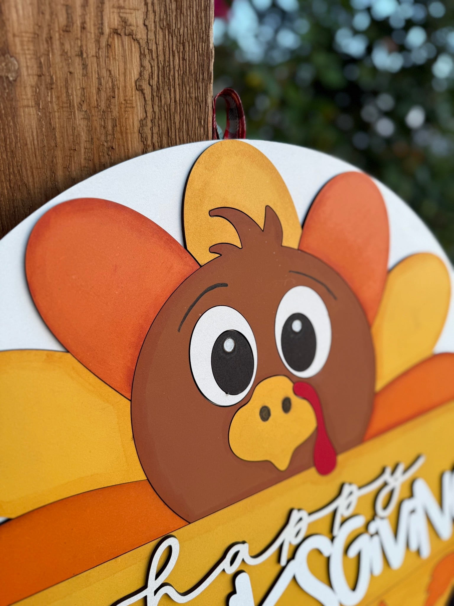 A close-up of a wooden Thanksgiving sign shaped like a cartoon turkey with colorful orange and yellow feathers, hanging on a post. The turkey has wide eyes and a red wattle. Greenery is blurred in the background.