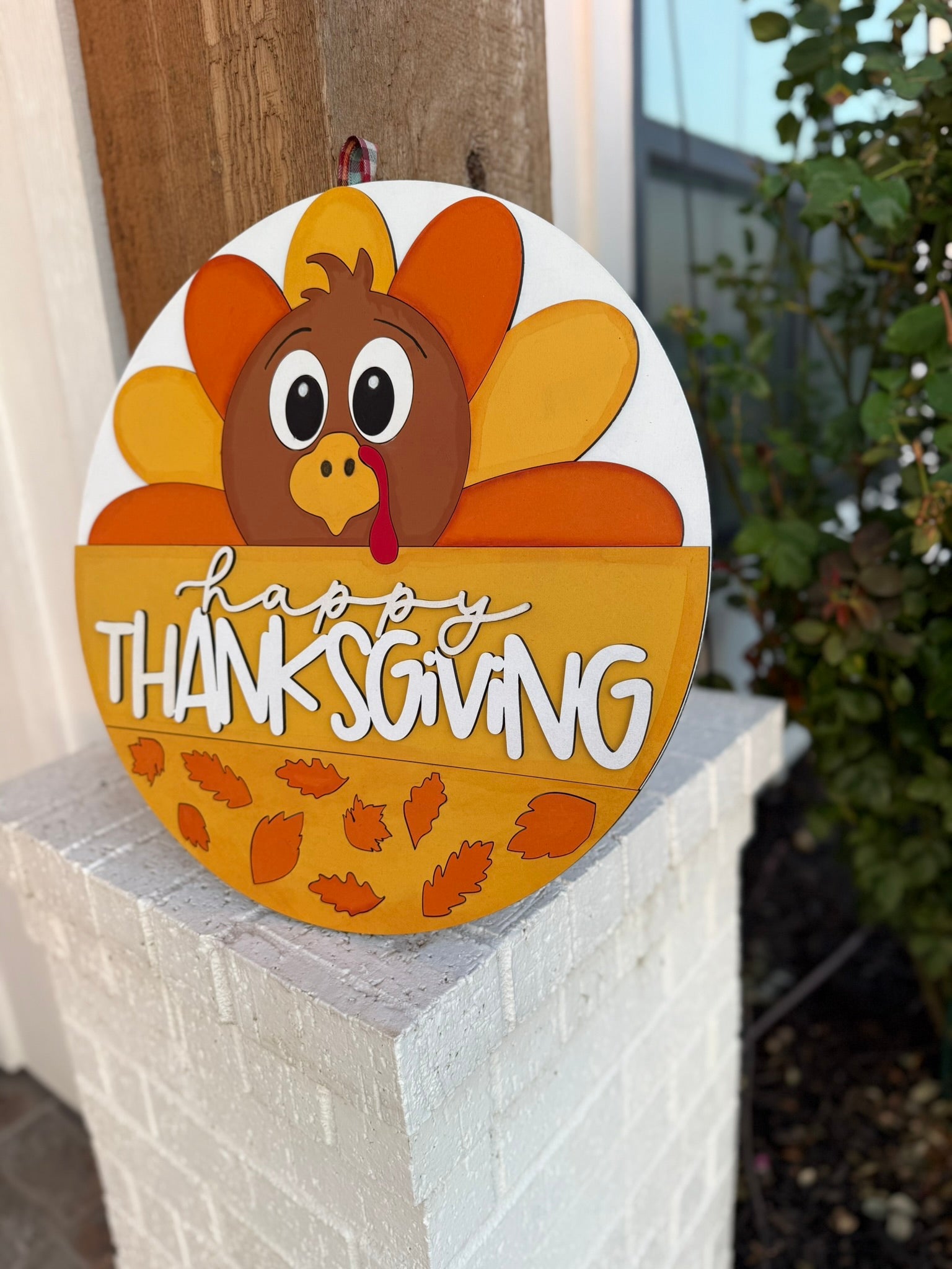 A round Happy Thanksgiving sign featuring a cartoon turkey with orange feathers, autumn leaves, and decorative lettering, hanging on a white brick post near a leafy bush.