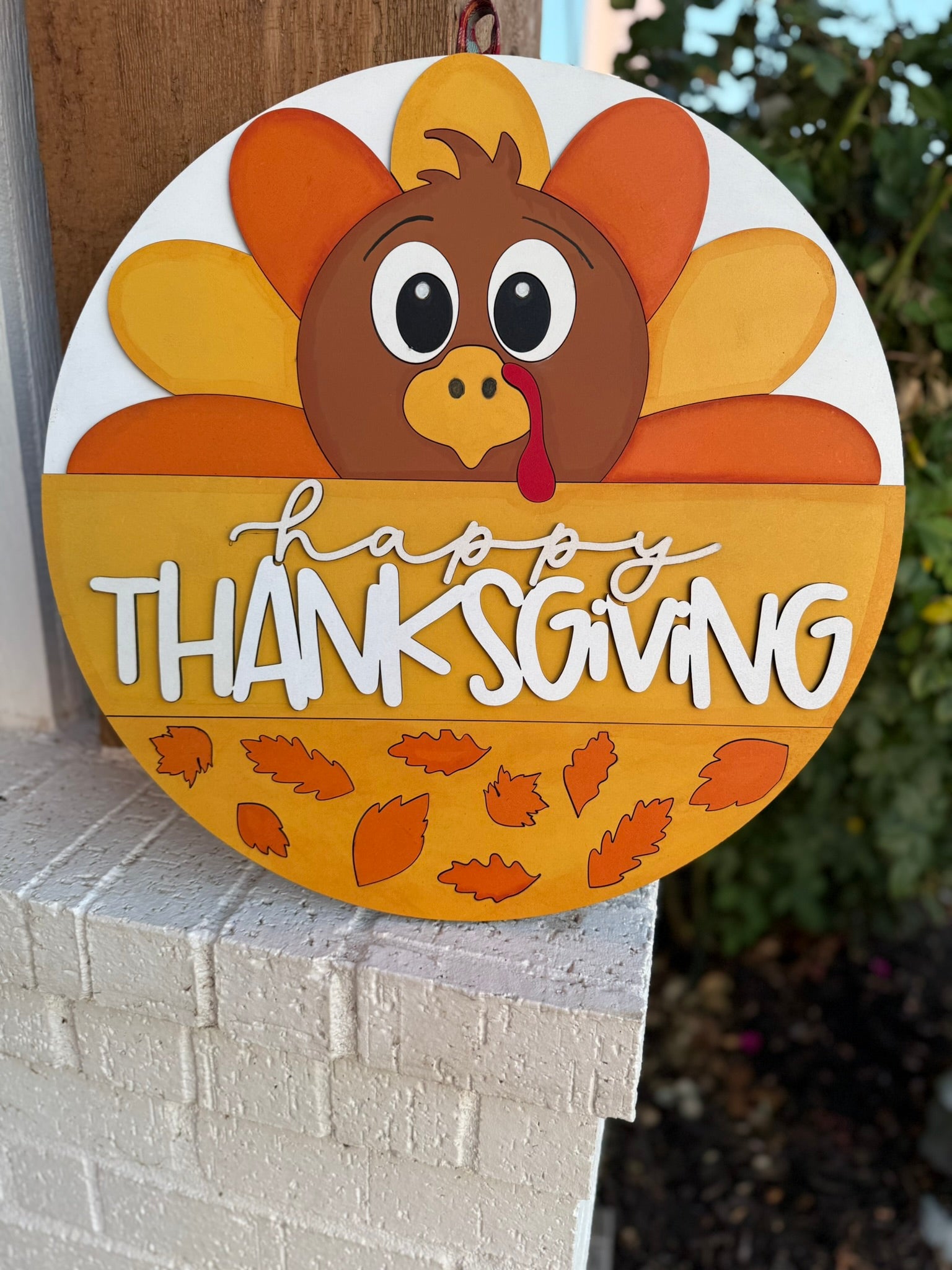 A round sign featuring a cartoon turkey with colorful feathers and the words happy THANKSGIVING in white letters, along with autumn leaves at the bottom, hangs on a white brick wall.