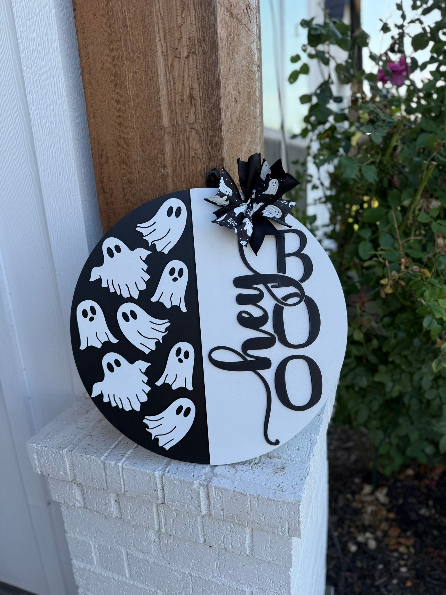 A round black-and-white sign leans against a wall, decorated with cartoon ghosts on one side and the words hey BOO in large letters on the other, topped with a black-and-white bow. Roses and greenery are in the background.