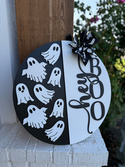 A round wooden sign split black and white, decorated with white ghost cutouts on the black half and the words hey BOO on the white half, topped with a black ribbon bow featuring ghost designs.