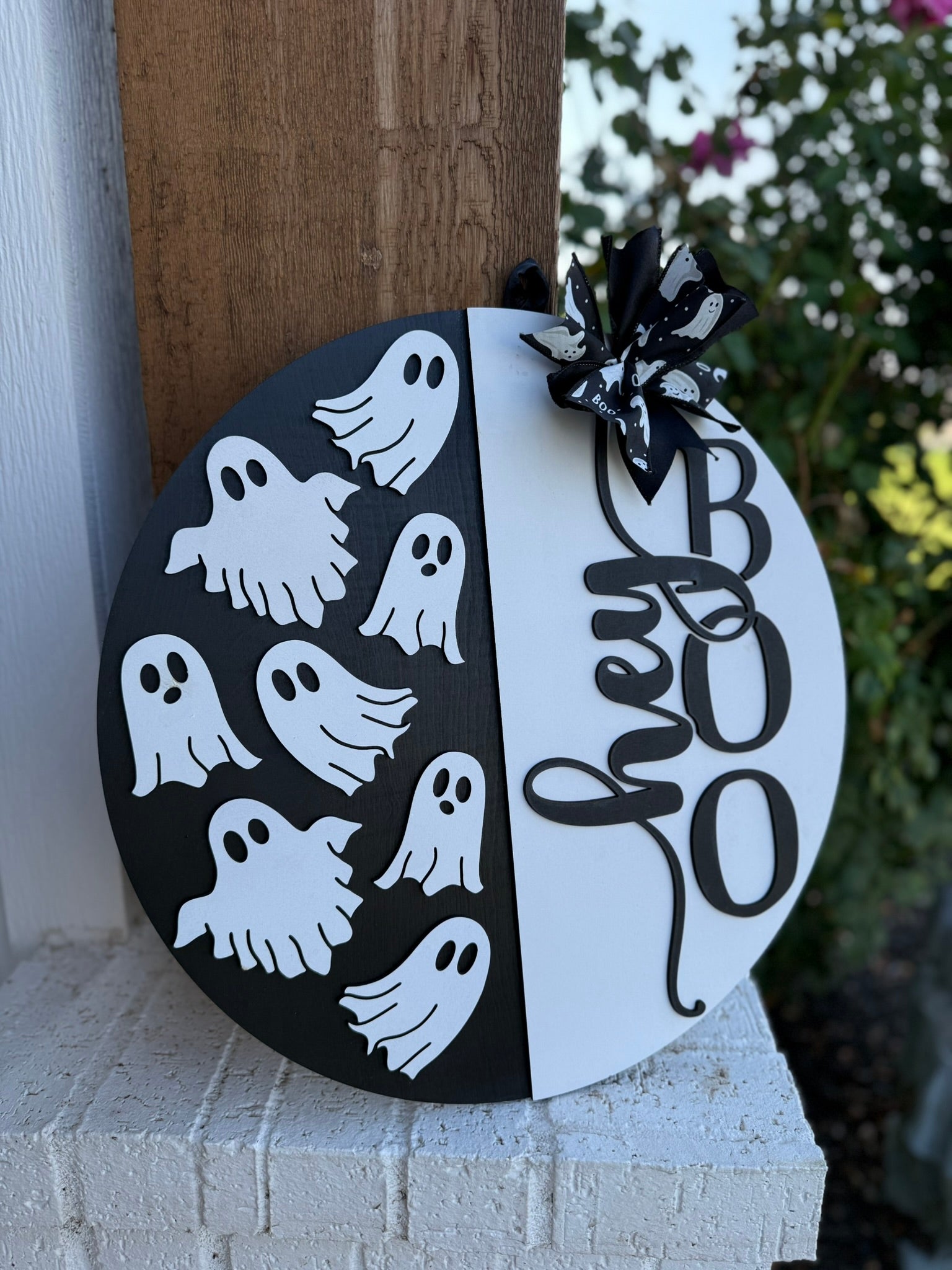 A round sign with a black-and-white design reads “hey BOO” in large letters and features several cartoon ghost shapes and a black-and-white bow on top. The sign is displayed outside on a white column.