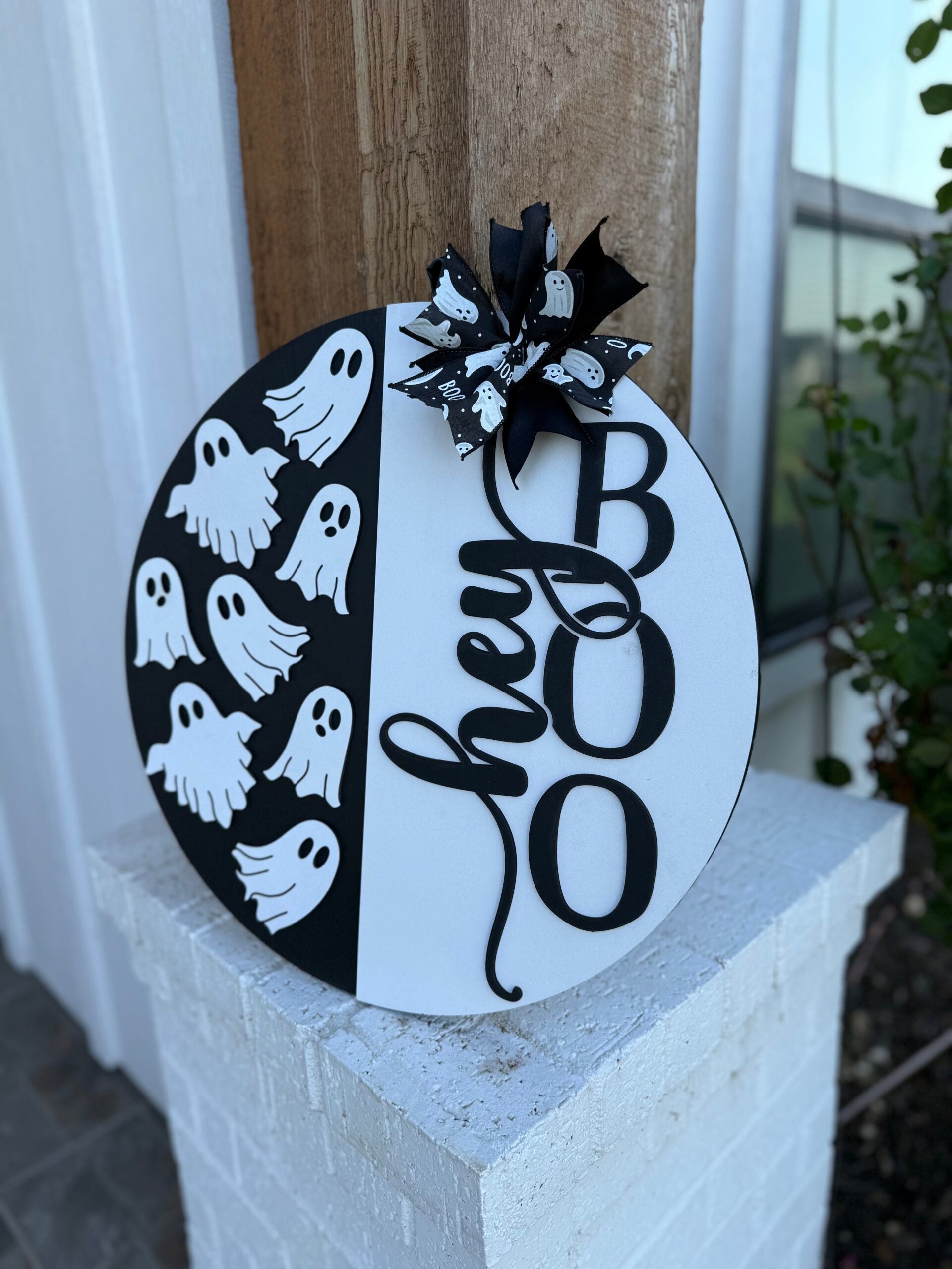 A round black and white sign with “hey BOO” written in large letters and decorated with a bow and cartoon ghosts, displayed on a porch pillar near a house.
