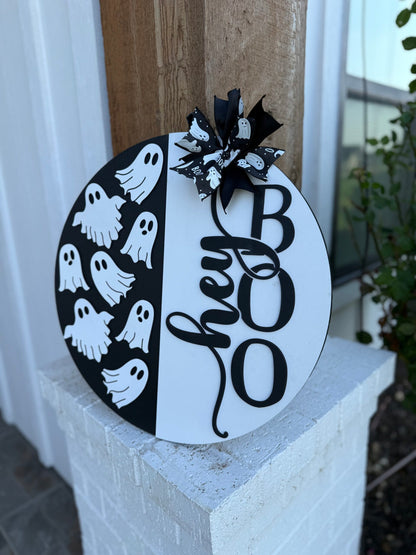 A round black and white sign with “hey BOO” written in large letters and decorated with a bow and cartoon ghosts, displayed on a porch pillar near a house.