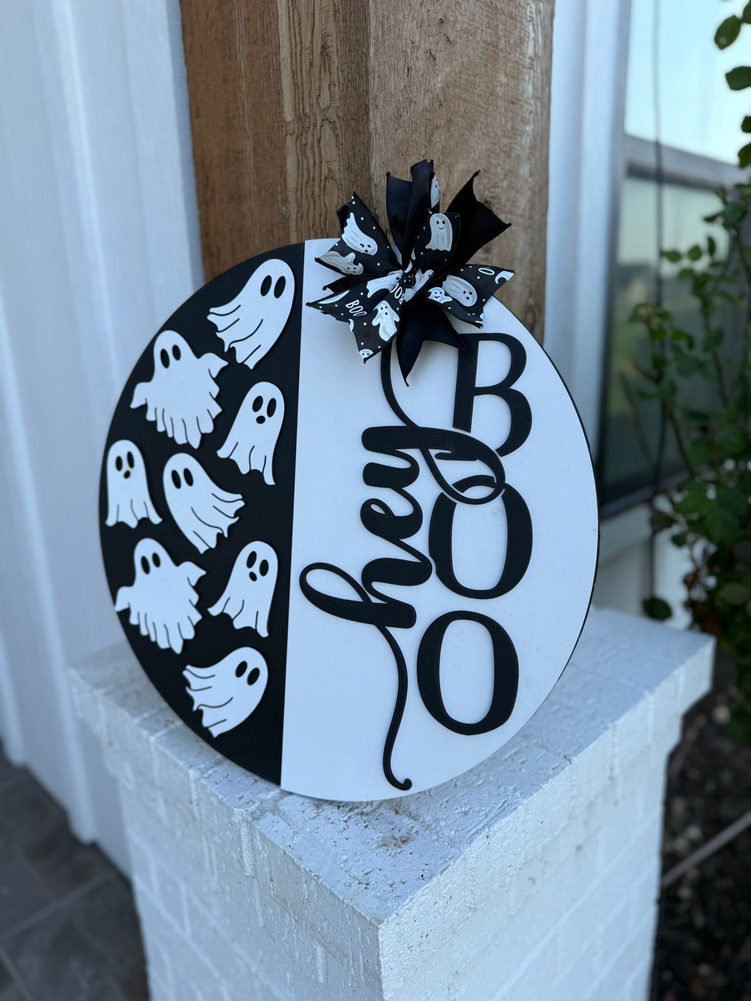 A round sign with a black-and-white design reads “hey BOO” in large letters and features several cartoon ghost shapes and a black-and-white bow on top. The sign is displayed outside on a white column.