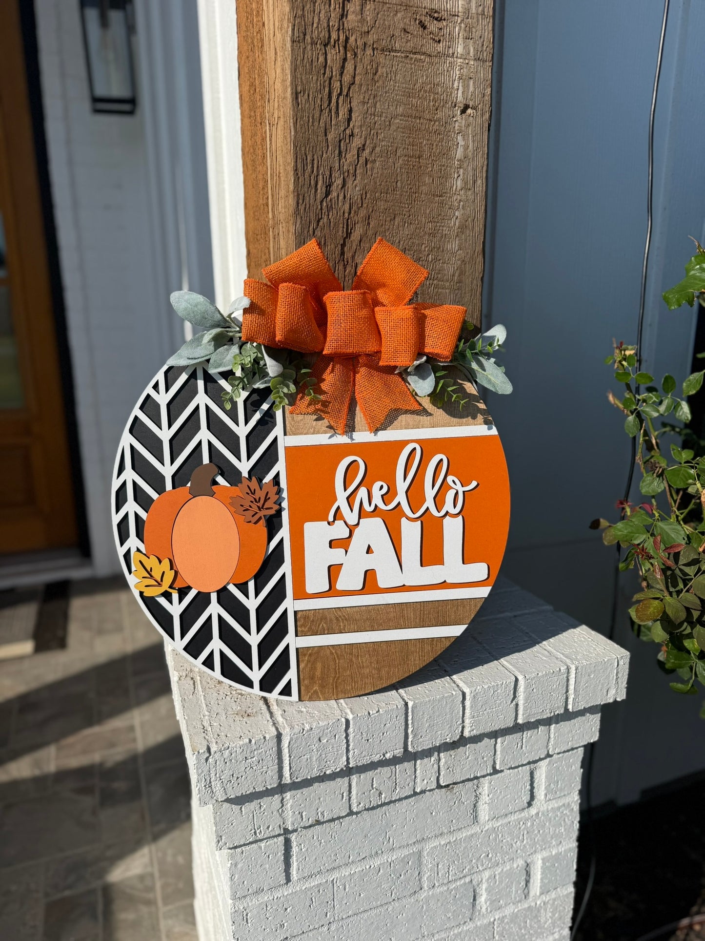 A round wooden sign decorated with orange pumpkins, leaves, greenery, and a large orange bow reads hello FALL in white and orange letters, displayed on a porch post in bright sunlight.