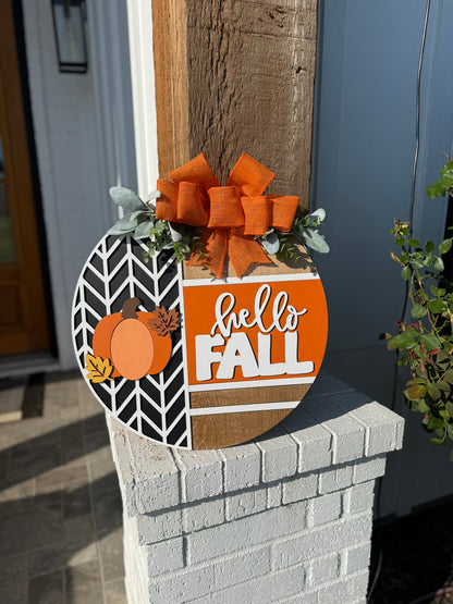 A round wooden sign decorated with orange pumpkins, leaves, greenery, and a large orange bow reads hello FALL in white and orange letters, displayed on a porch post in bright sunlight.
