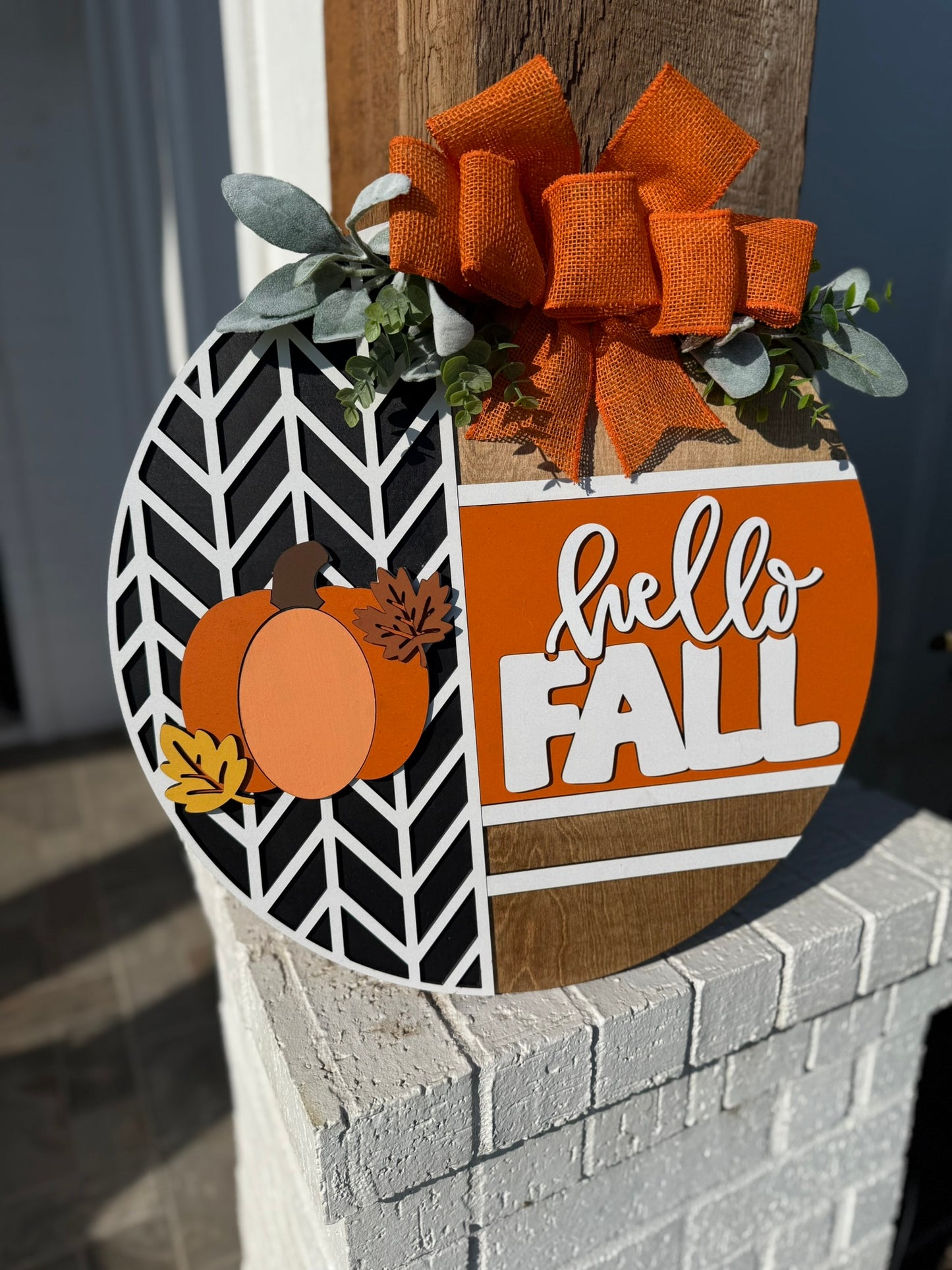 A round wooden sign decorated with faux greenery and an orange burlap bow, featuring a pumpkin design and the words “hello FALL” in white on an orange background. The sign is mounted on a post.