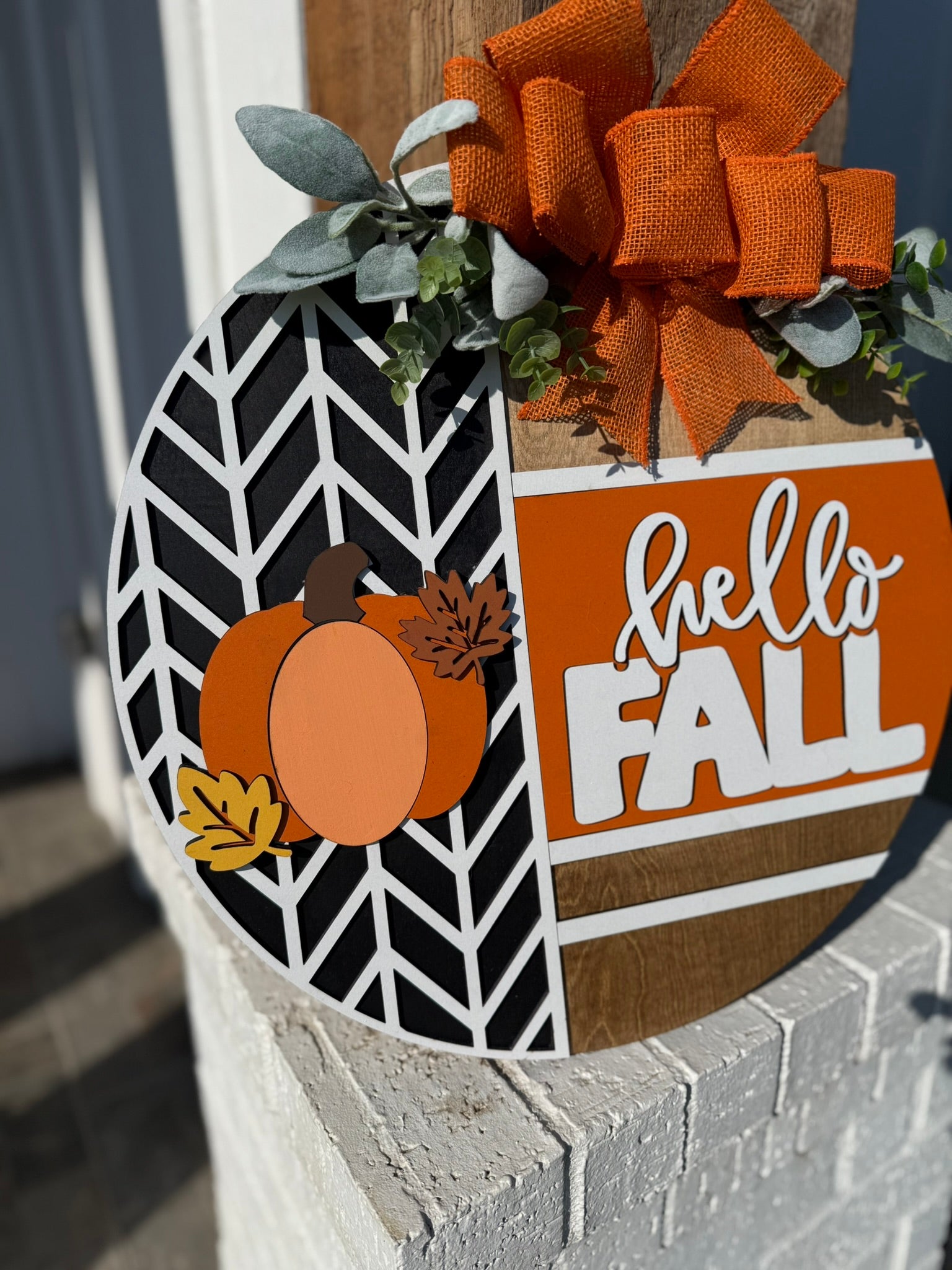 A round decorative sign features a pumpkin, leaves, and the words hello FALL in white and orange. It is adorned with a large orange bow and greenery, and hangs on a white brick post.