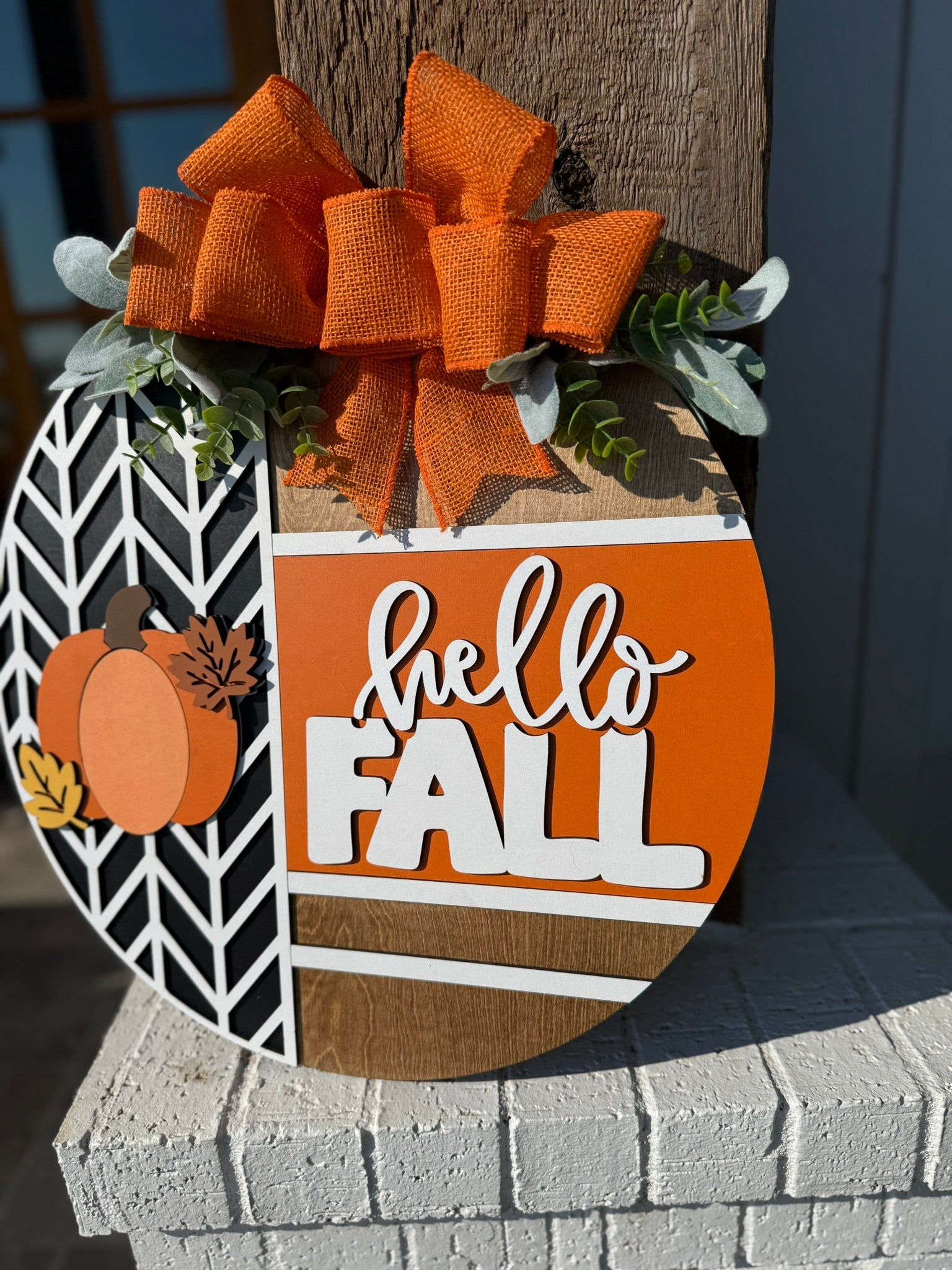 A circular wooden sign with an orange bow, greenery, and the words hello FALL in white letters, decorated with pumpkins and leaves, hangs on a porch pillar.