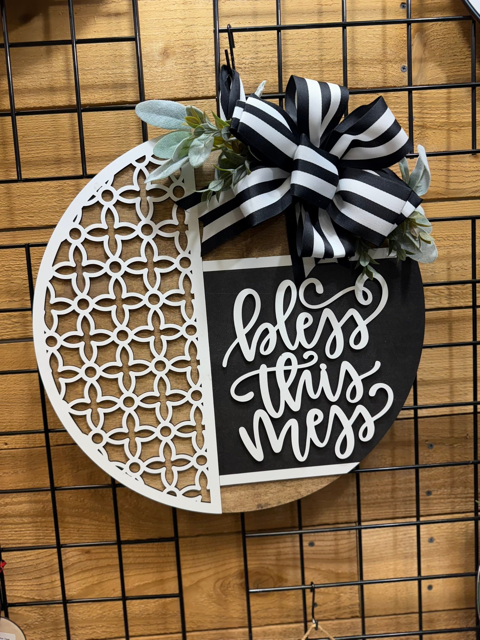 Round wall sign with a black-and-white floral pattern on the left, a striped black-and-white bow and greenery on top, and the phrase “bless this mess” written in white script on the right.