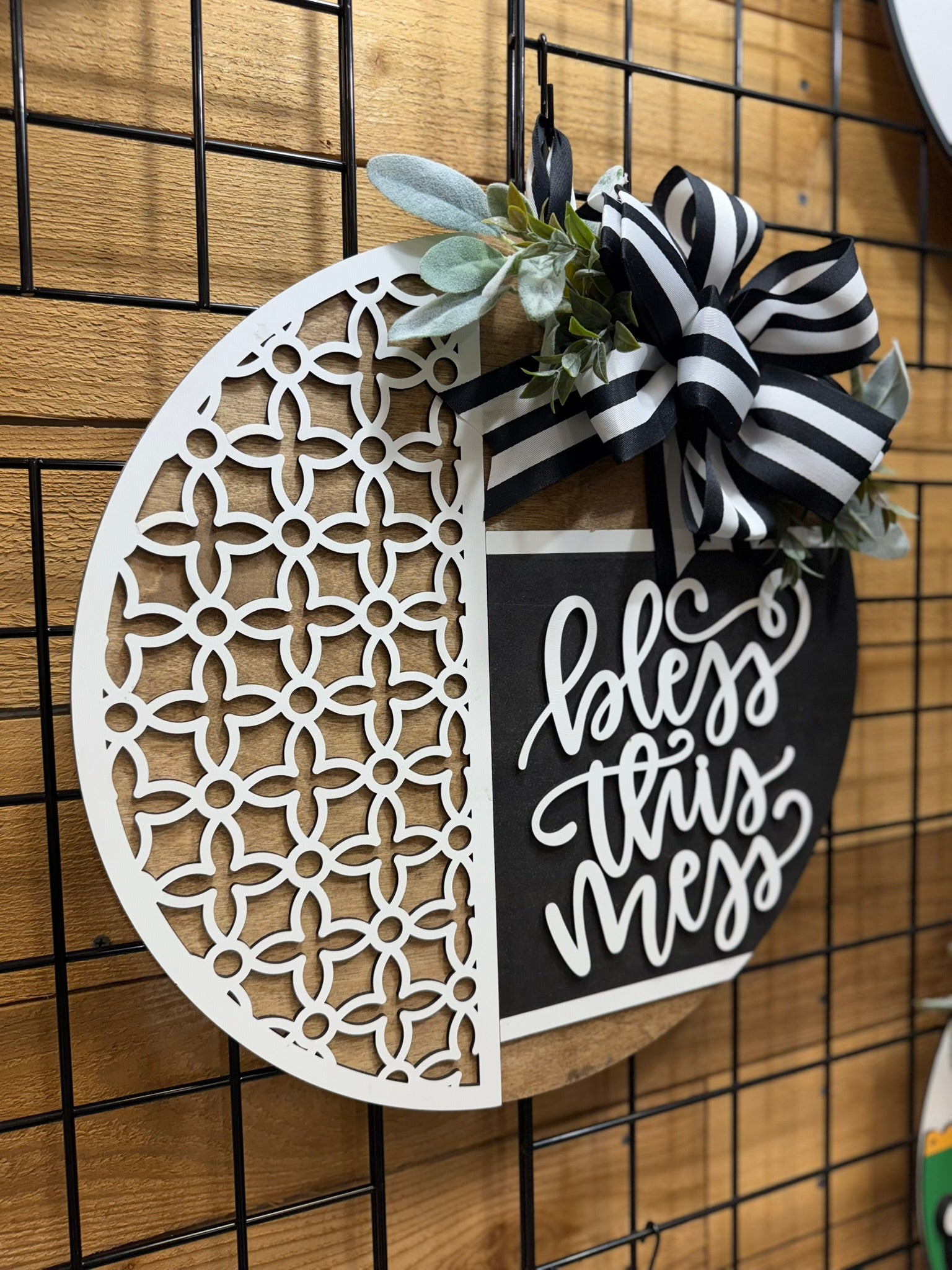 A round wooden sign with a white decorative lattice on the left, greenery and a black-and-white striped bow on top, and the phrase “bless this mess” in white script on a black background on the right.