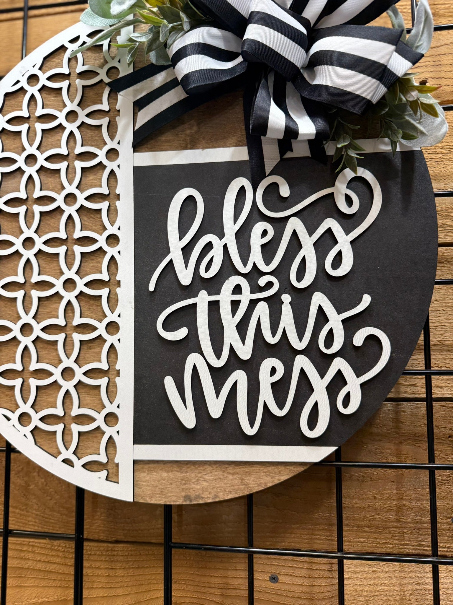 A decorative round sign with a patterned white cutout on the left and the words bless this mess in white script on a black background, topped with a black and white striped bow and greenery.