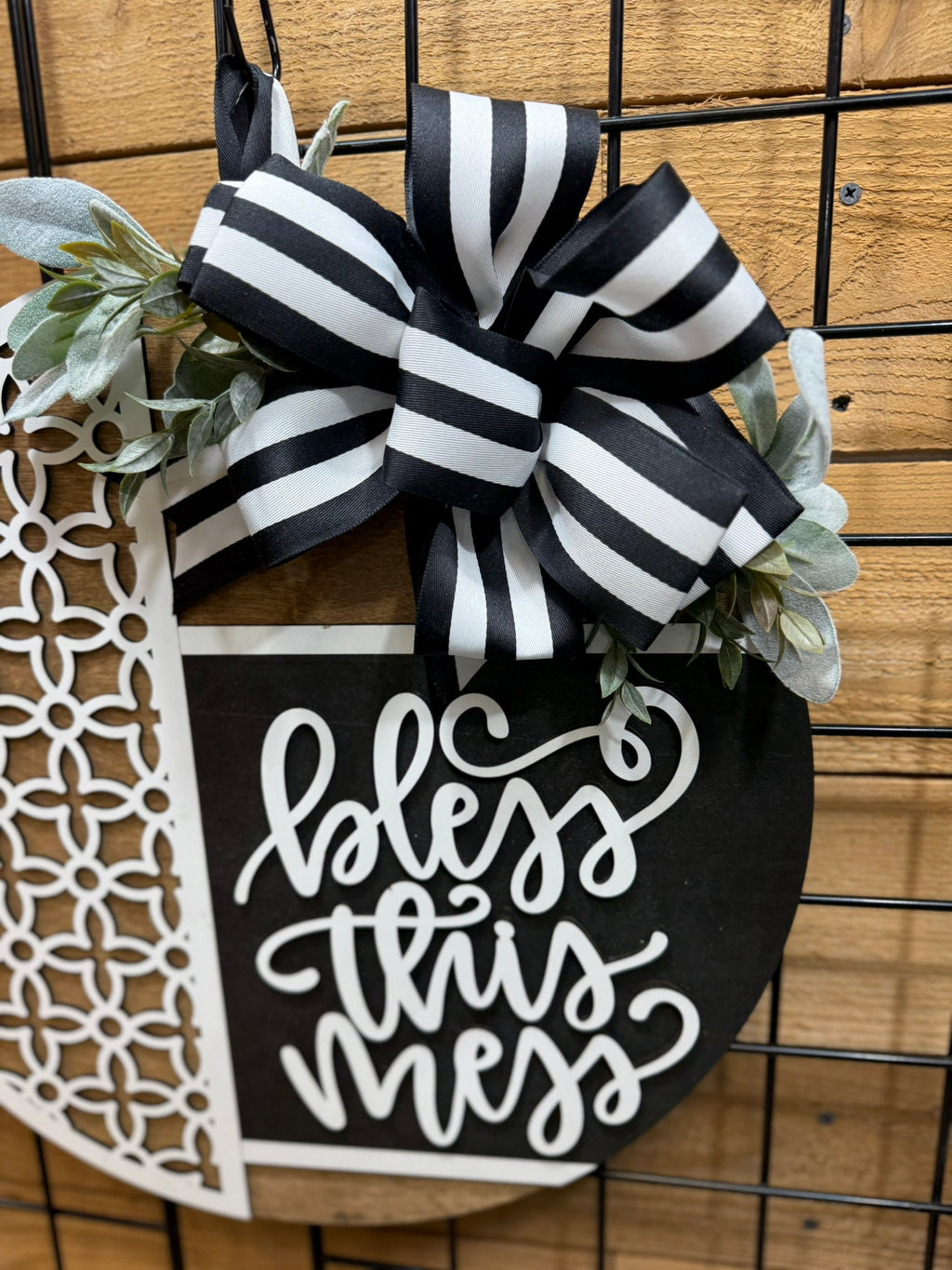 A black and white circular sign with a striped bow and greenery reads bless this mess in cursive, hanging on a wooden and metal grid background.