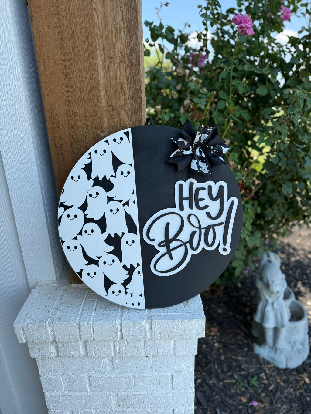 A round black and white Halloween sign features cartoon ghosts on one side, a Hey Boo! message in bold script on the other, and a bow with ghost patterns at the top. The sign is propped against a white brick wall.