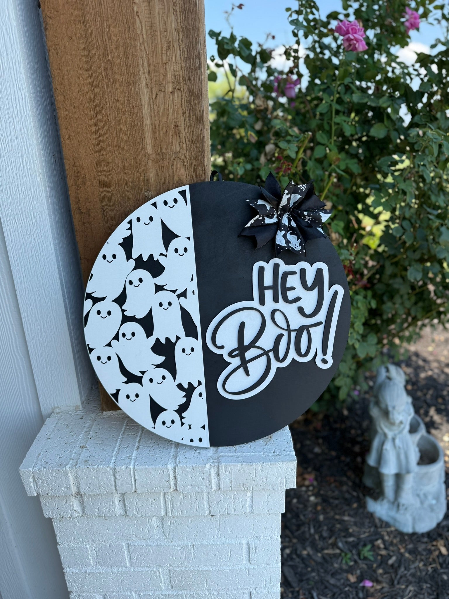 A round black and white Halloween sign features cartoon ghosts on one side, a Hey Boo! message in bold script on the other, and a bow with ghost patterns at the top. The sign is propped against a white brick wall.