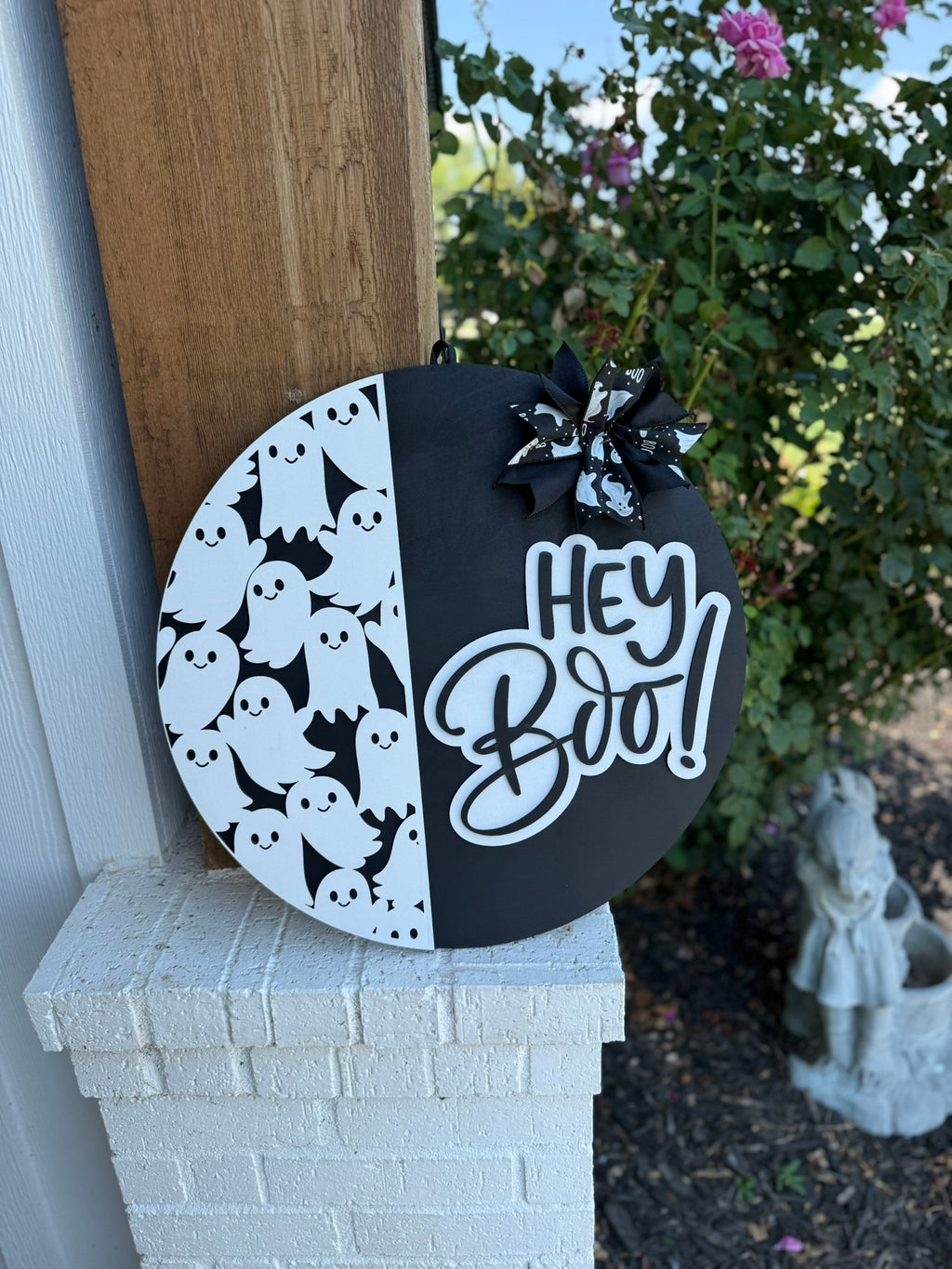 A round black and white Halloween sign features cartoon ghosts on one side, a Hey Boo! message in bold script on the other, and a bow with ghost patterns at the top. The sign is propped against a white brick wall.