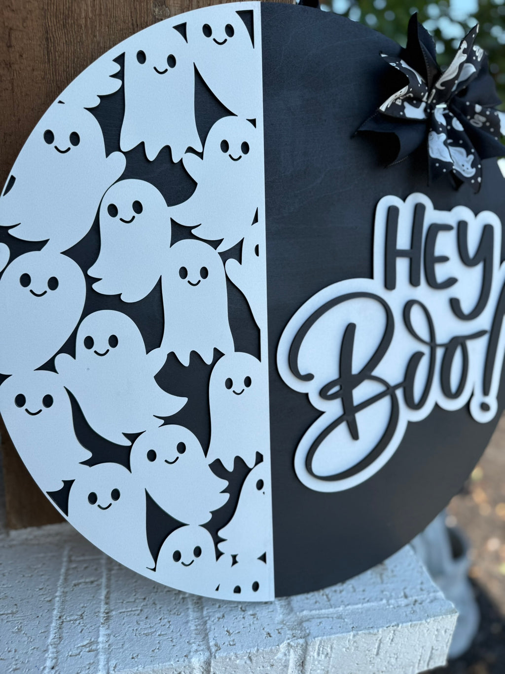A round black and white Halloween sign features cartoon ghosts on one side, a Hey Boo! message in bold script on the other, and a bow with ghost patterns at the top. The sign is propped against a white brick wall.