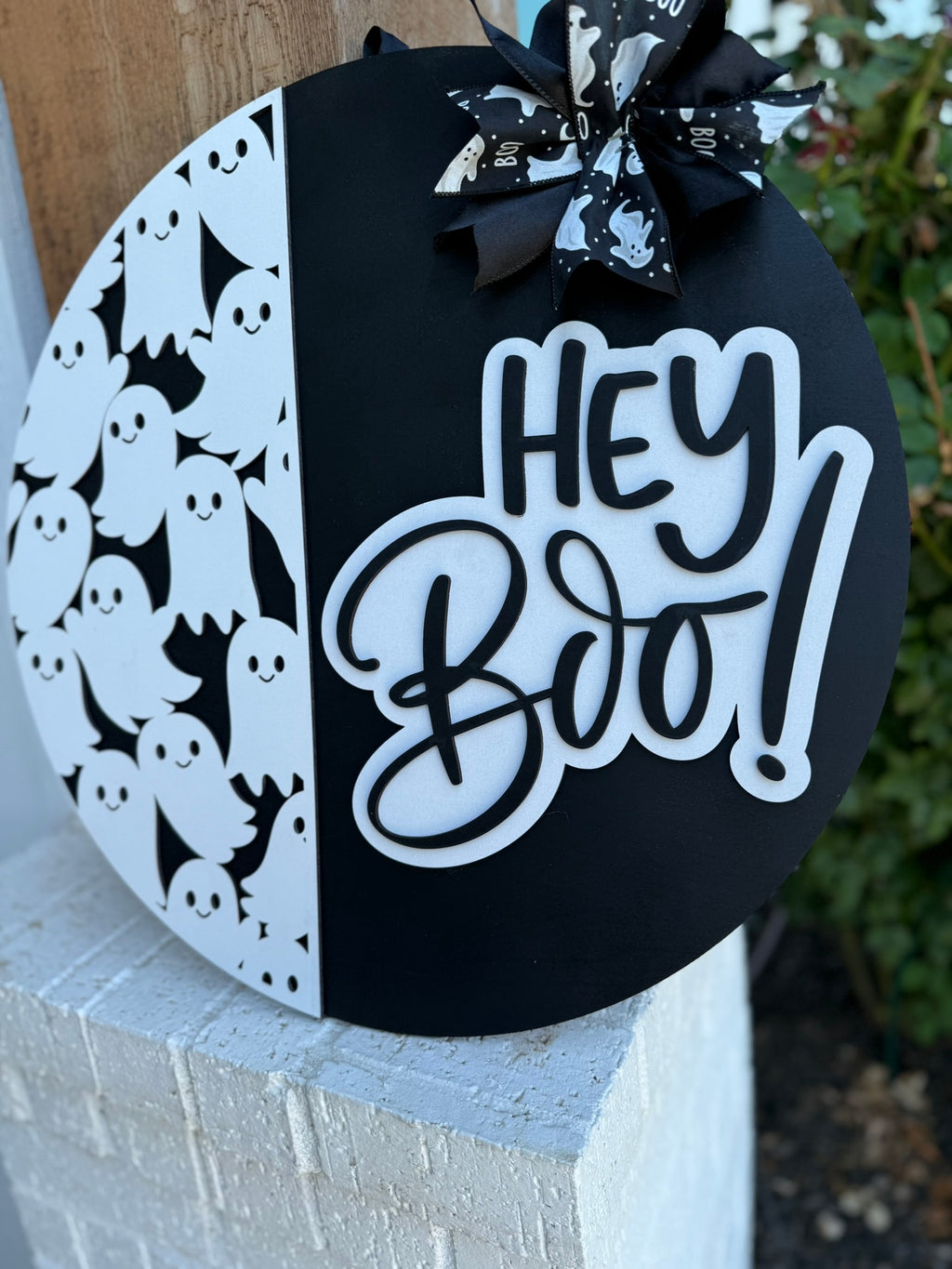 A round black and white Halloween sign features cartoon ghosts on one side, a Hey Boo! message in bold script on the other, and a bow with ghost patterns at the top. The sign is propped against a white brick wall.