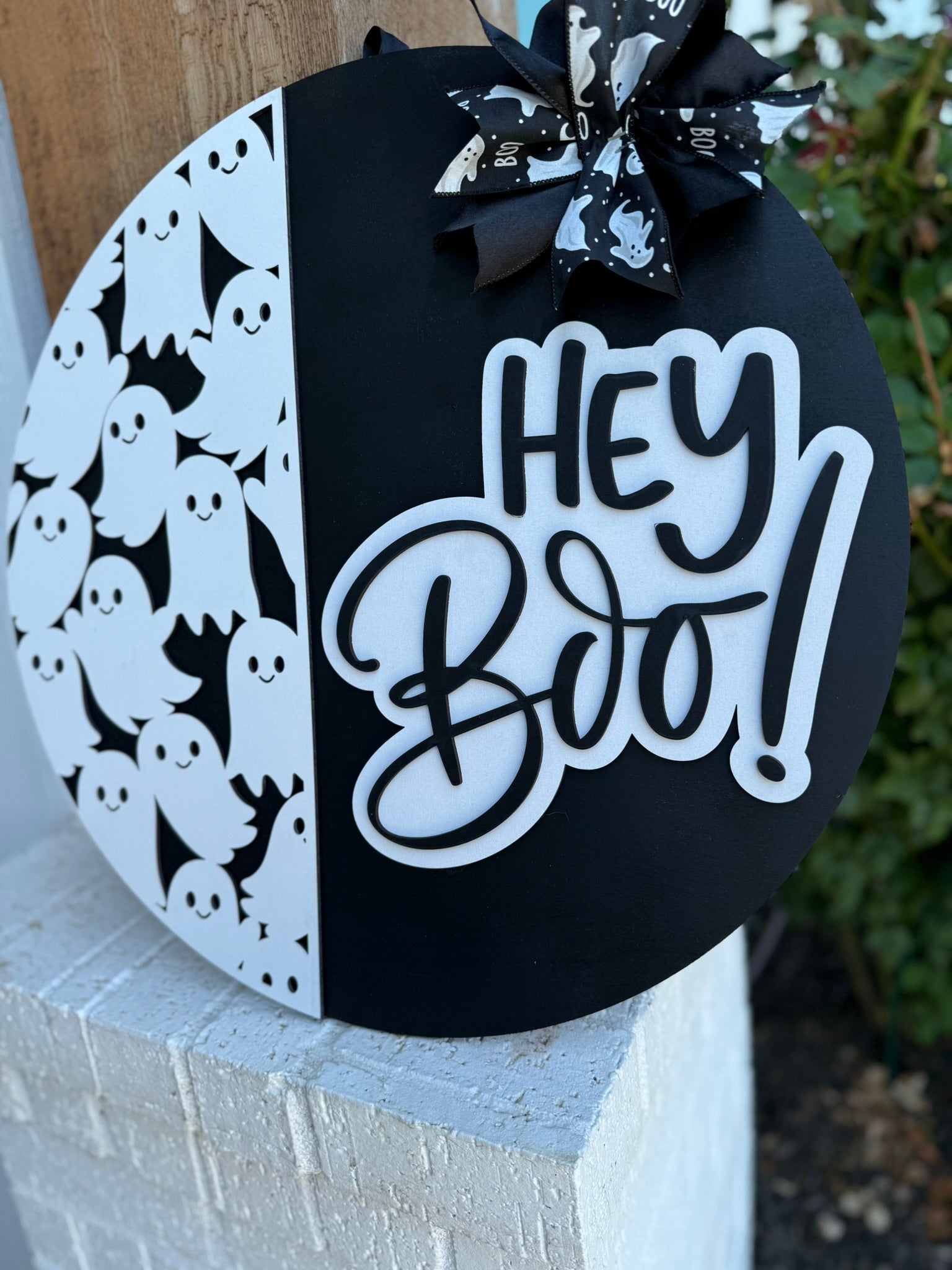 A round black and white Halloween sign features cartoon ghosts on one side, a Hey Boo! message in bold script on the other, and a bow with ghost patterns at the top. The sign is propped against a white brick wall.