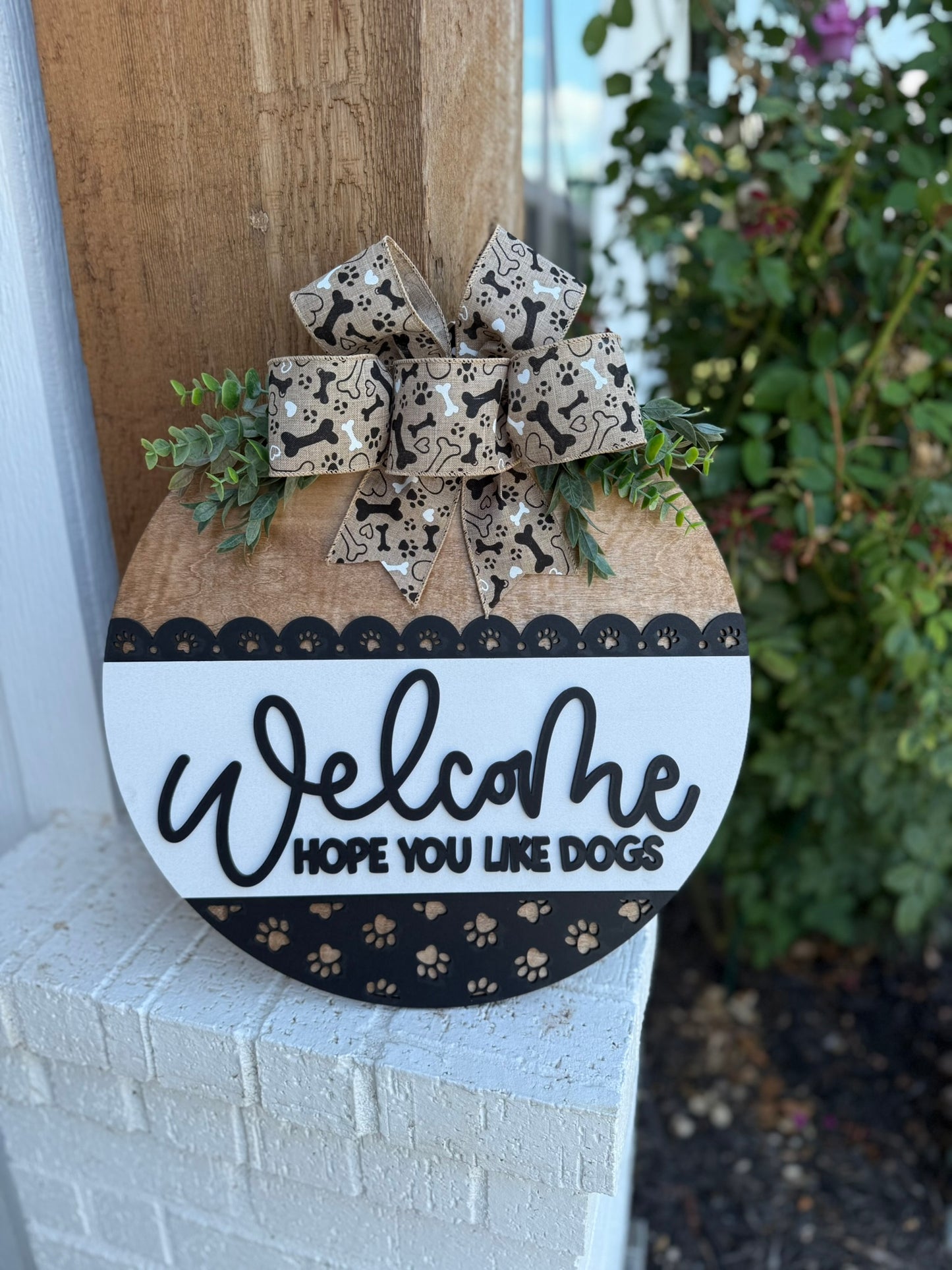 A smiling woman holding a round sign that says Welcome, hope you like dogs with a paw print design and a decorative bow, standing in front of a modern house.