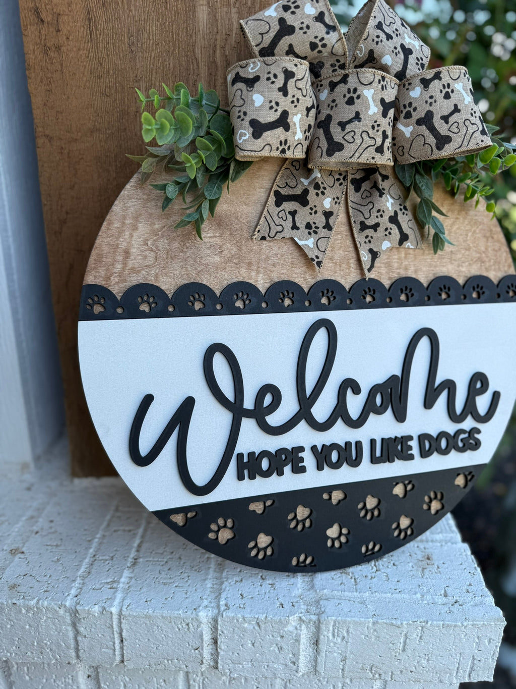 A smiling woman holding a round sign that says Welcome, hope you like dogs with a paw print design and a decorative bow, standing in front of a modern house.