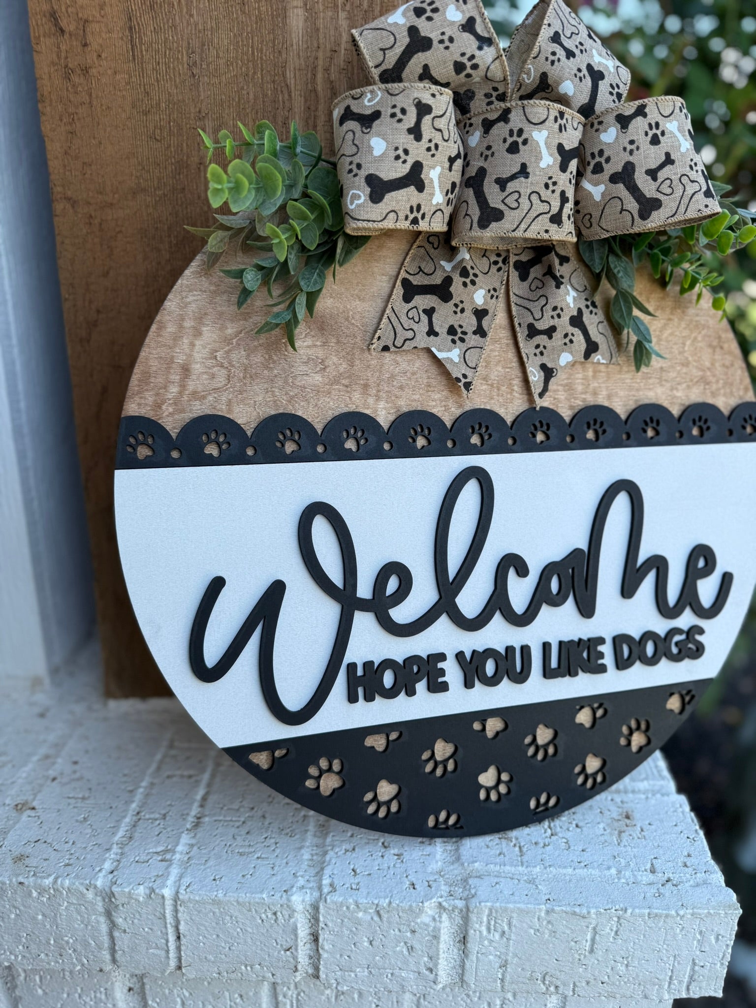 A smiling woman holding a round sign that says Welcome, hope you like dogs with a paw print design and a decorative bow, standing in front of a modern house.
