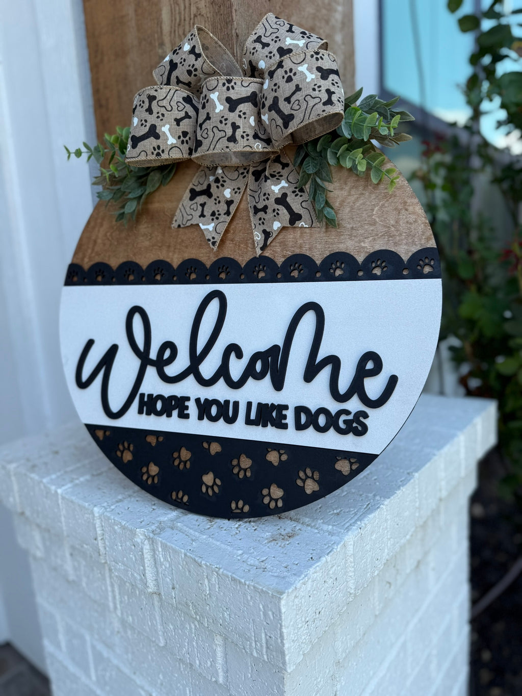 A smiling woman holding a round sign that says Welcome, hope you like dogs with a paw print design and a decorative bow, standing in front of a modern house.