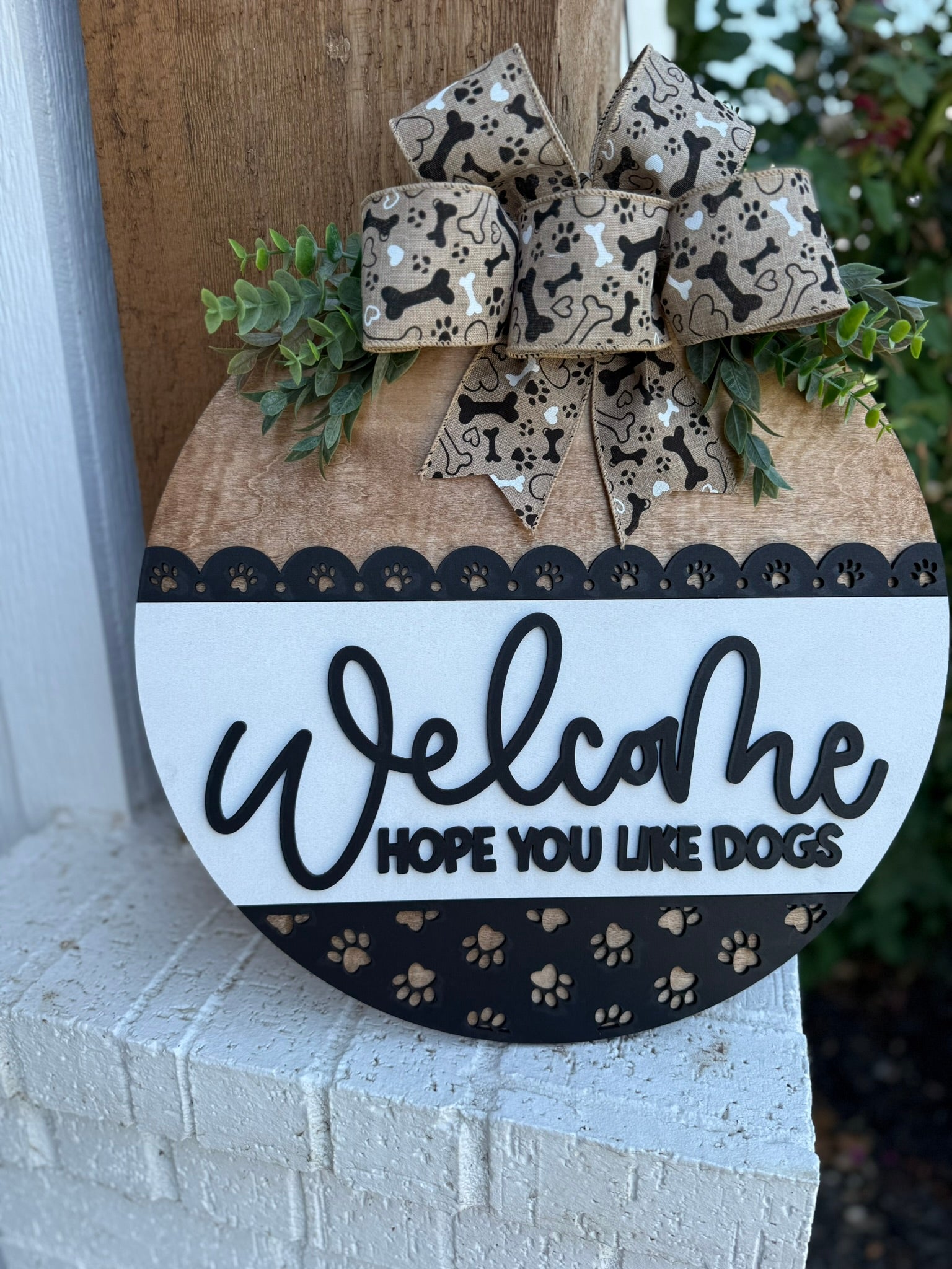 A smiling woman holding a round sign that says Welcome, hope you like dogs with a paw print design and a decorative bow, standing in front of a modern house.
