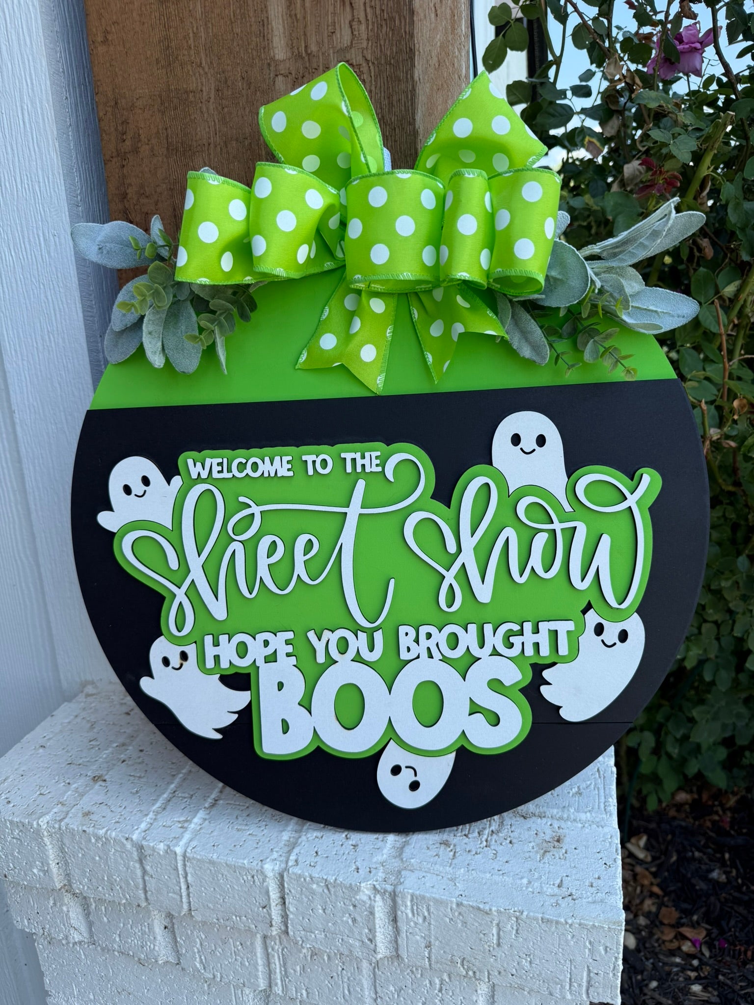 A round Halloween sign with a green top, polka-dot bow, leaves, and white ghosts, reading Welcome to the Sheet Show, hope you brought boos, displayed on a brick surface.