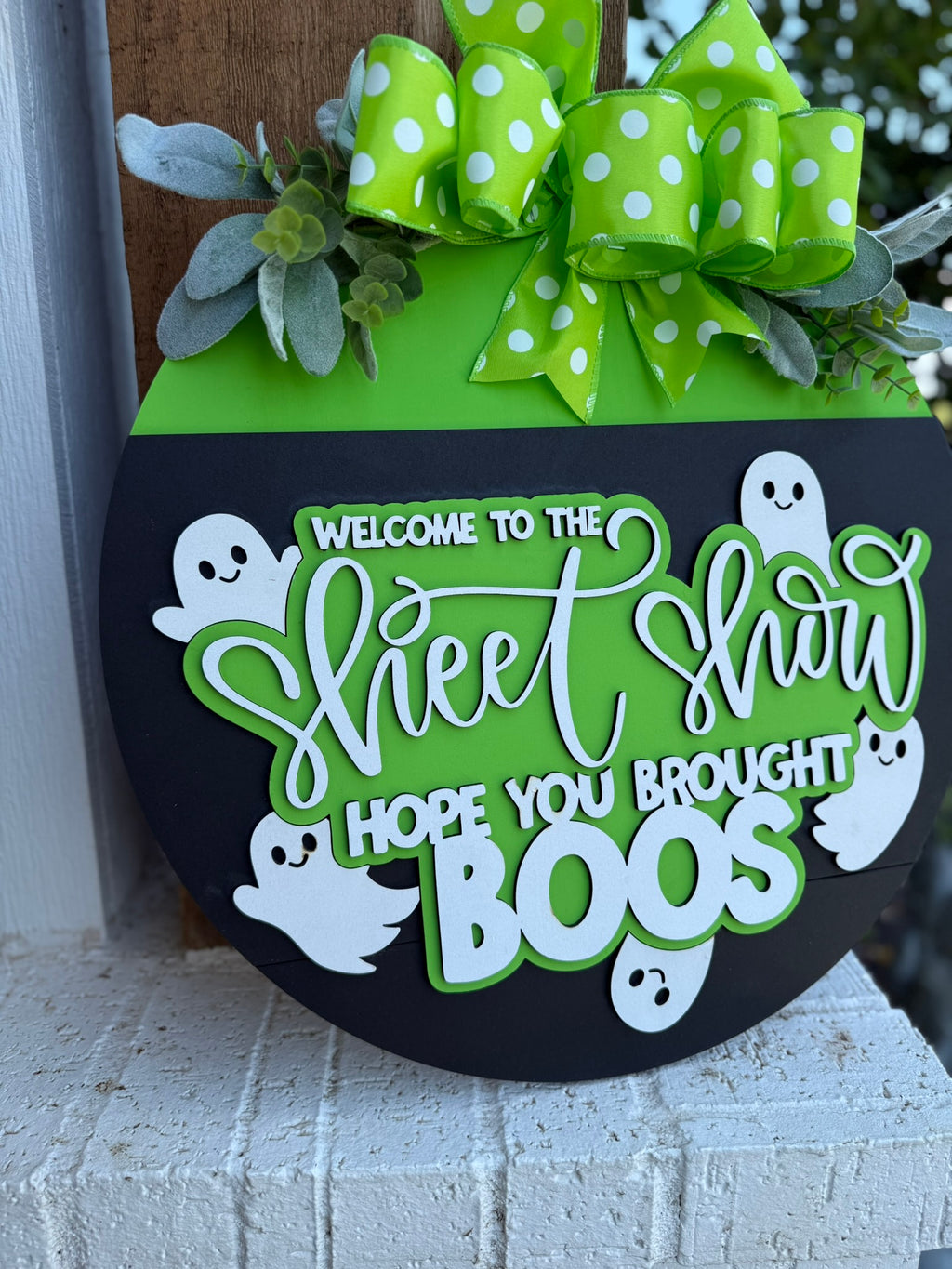 A round Halloween sign with a green top, polka-dot bow, leaves, and white ghosts, reading Welcome to the Sheet Show, hope you brought boos, displayed on a brick surface.