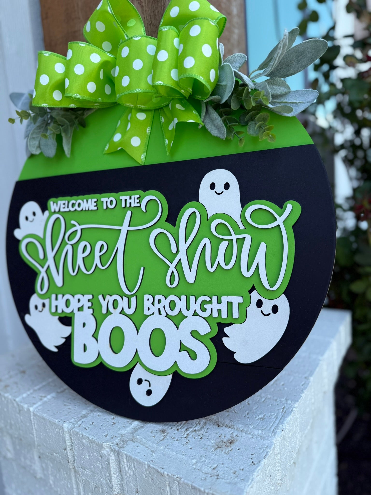 A round Halloween sign with a green top, polka-dot bow, leaves, and white ghosts, reading Welcome to the Sheet Show, hope you brought boos, displayed on a brick surface.