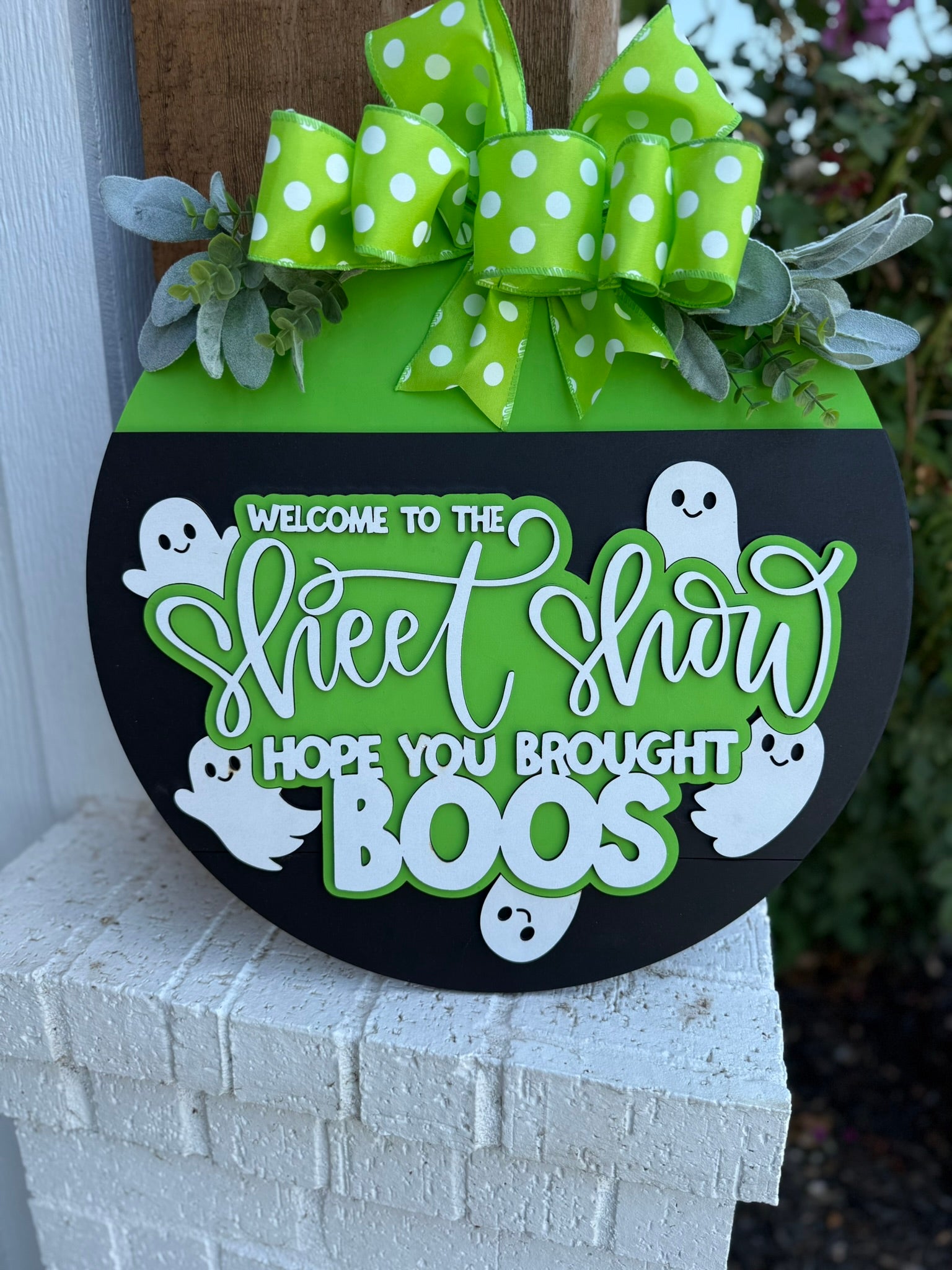 A round Halloween sign with a green top, polka-dot bow, leaves, and white ghosts, reading Welcome to the Sheet Show, hope you brought boos, displayed on a brick surface.