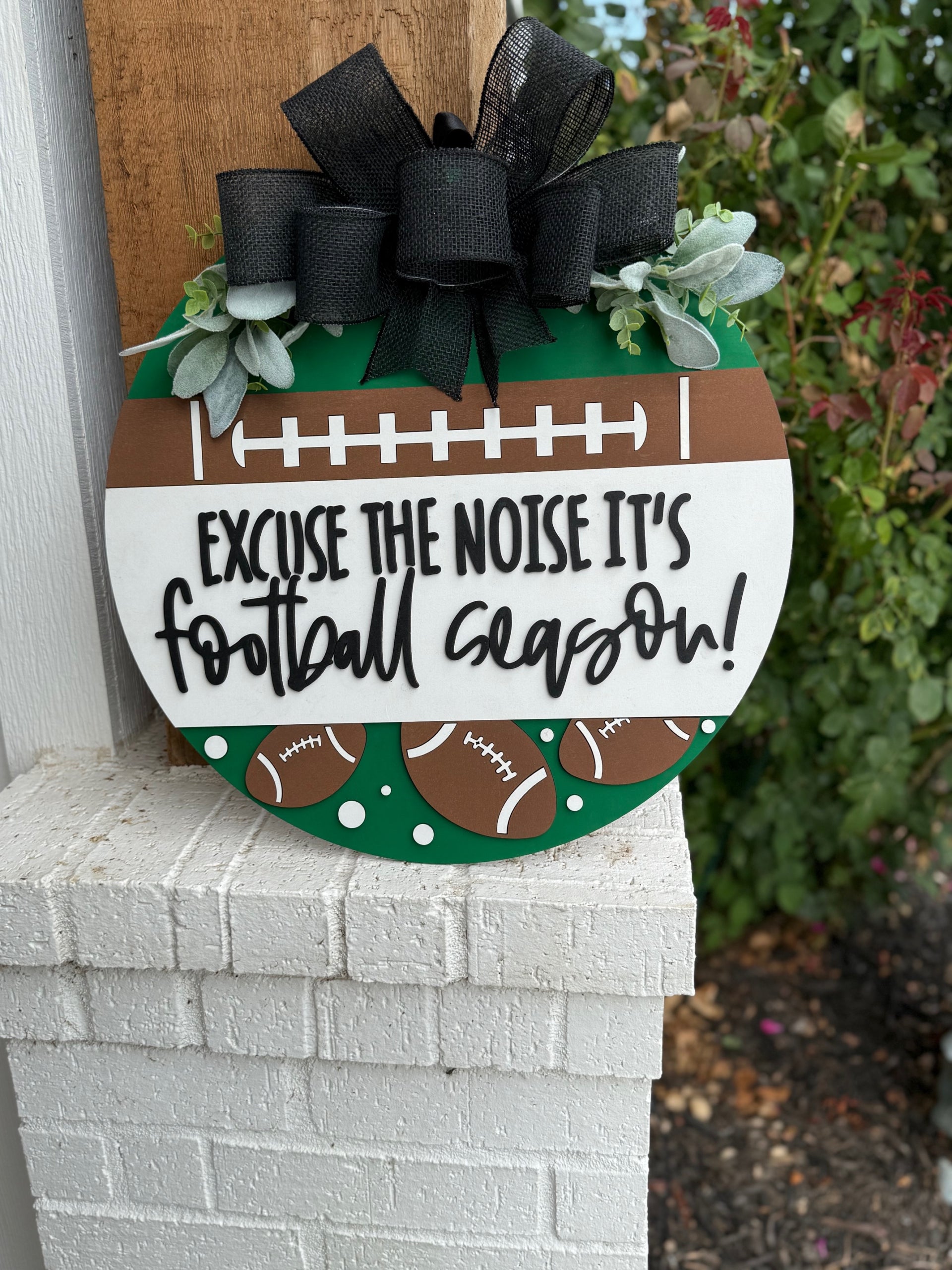 A round sign decorated with faux greenery and footballs reads, “Excuse the noise it’s football season” in bold, playful lettering. The sign features a football field design and is displayed against a white textured surface.