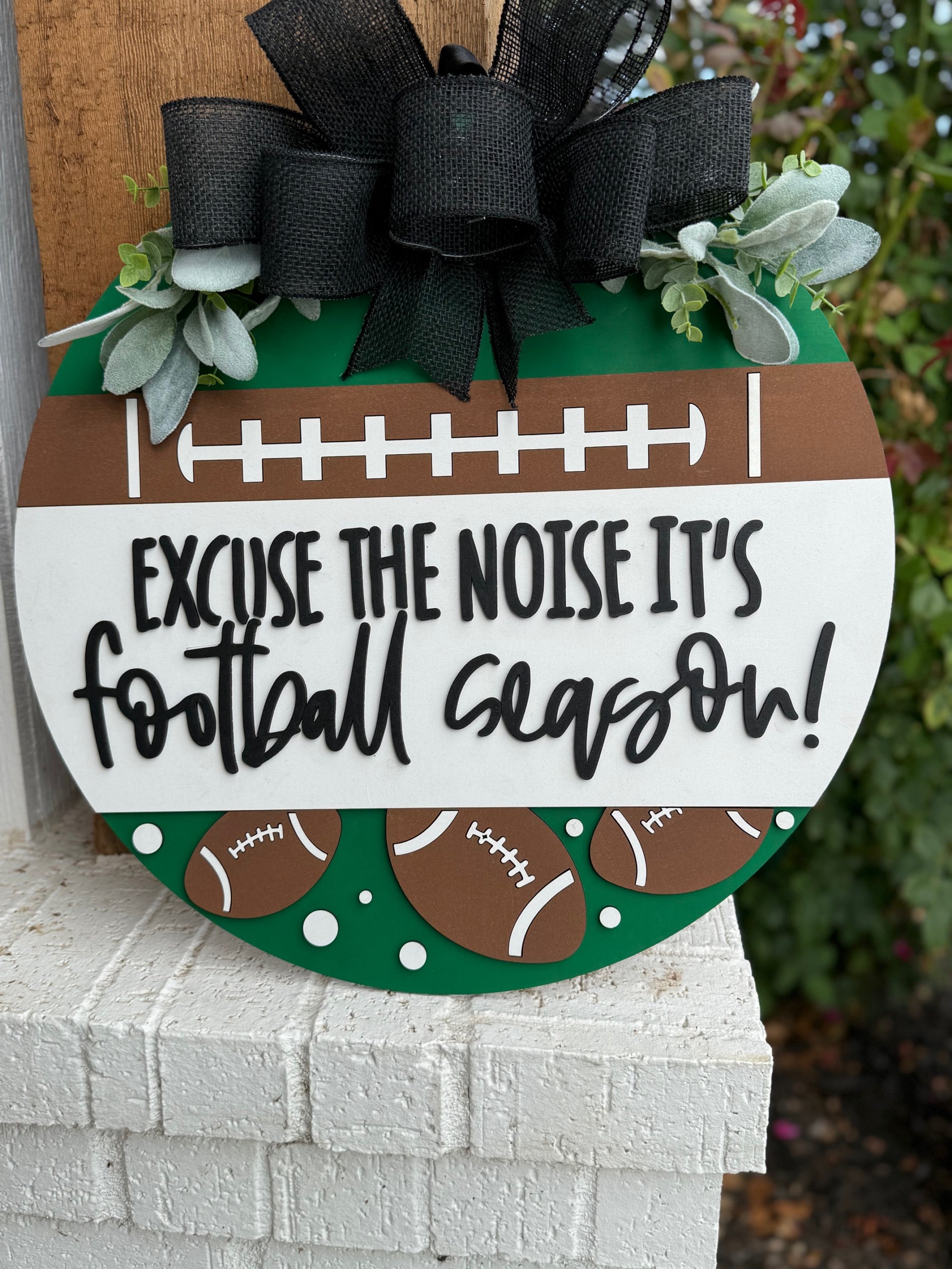 A round sign decorated with faux greenery and footballs reads, “Excuse the noise it’s football season” in bold, playful lettering. The sign features a football field design and is displayed against a white textured surface.