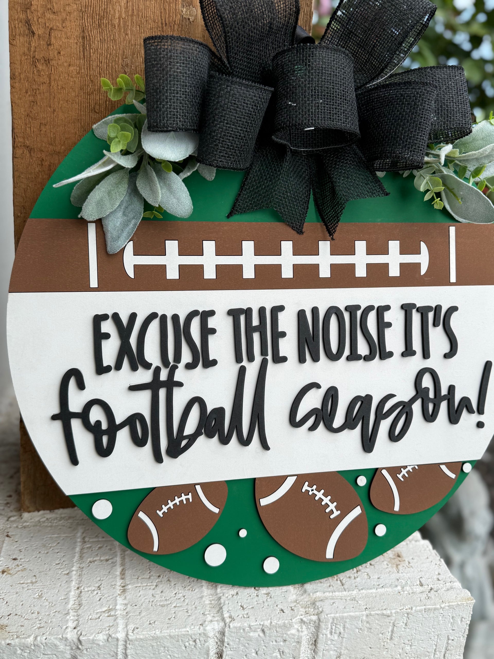 A round sign decorated with faux greenery and footballs reads, “Excuse the noise it’s football season” in bold, playful lettering. The sign features a football field design and is displayed against a white textured surface.