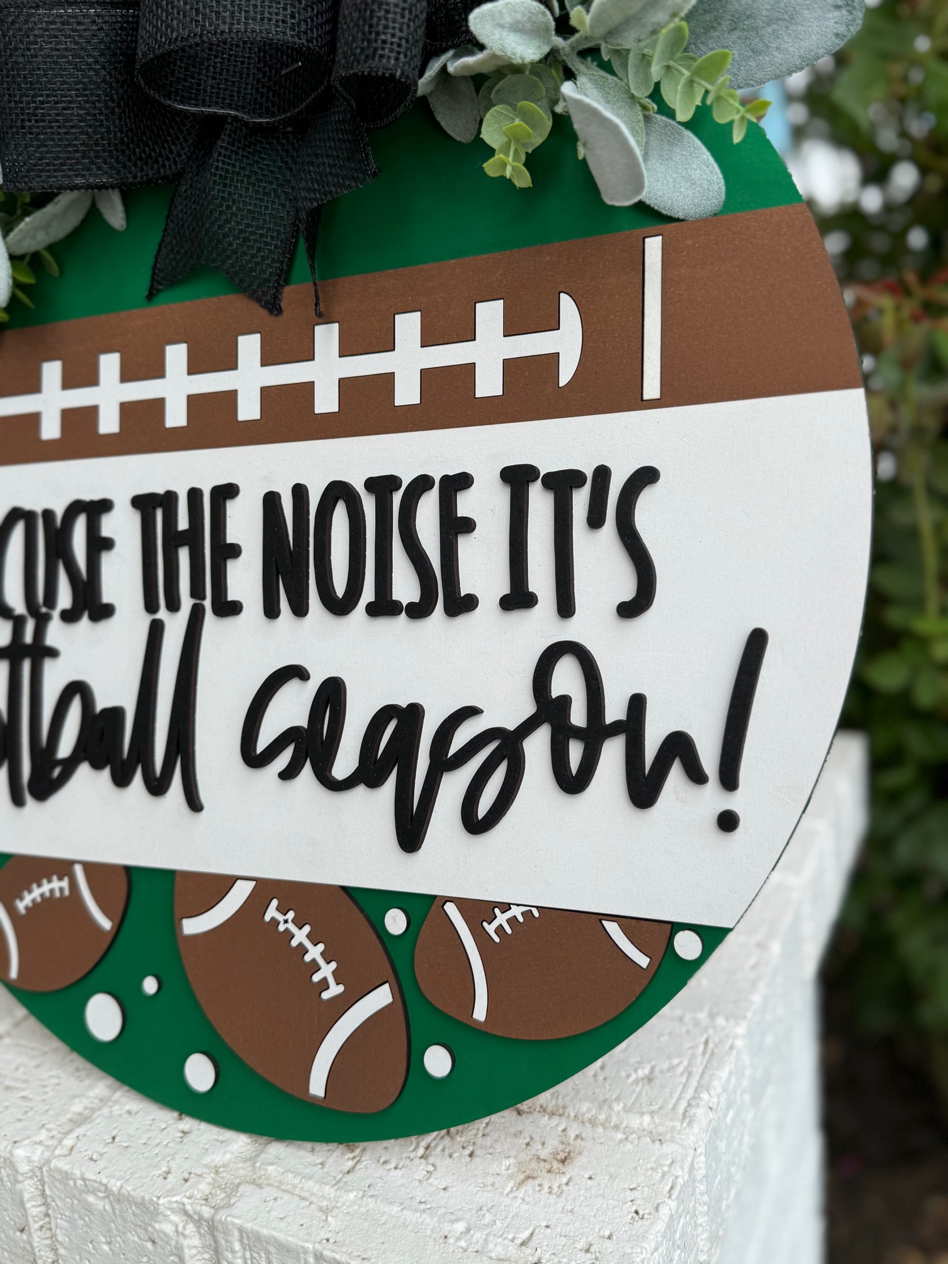 A round sign decorated with faux greenery and footballs reads, “Excuse the noise it’s football season” in bold, playful lettering. The sign features a football field design and is displayed against a white textured surface.