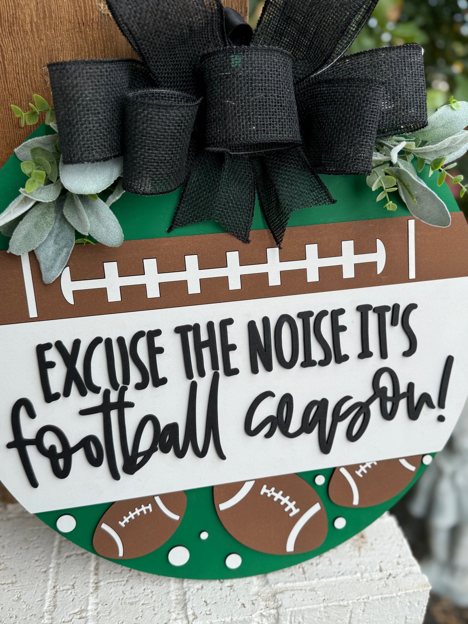 A round sign decorated with faux greenery and footballs reads, “Excuse the noise it’s football season” in bold, playful lettering. The sign features a football field design and is displayed against a white textured surface.