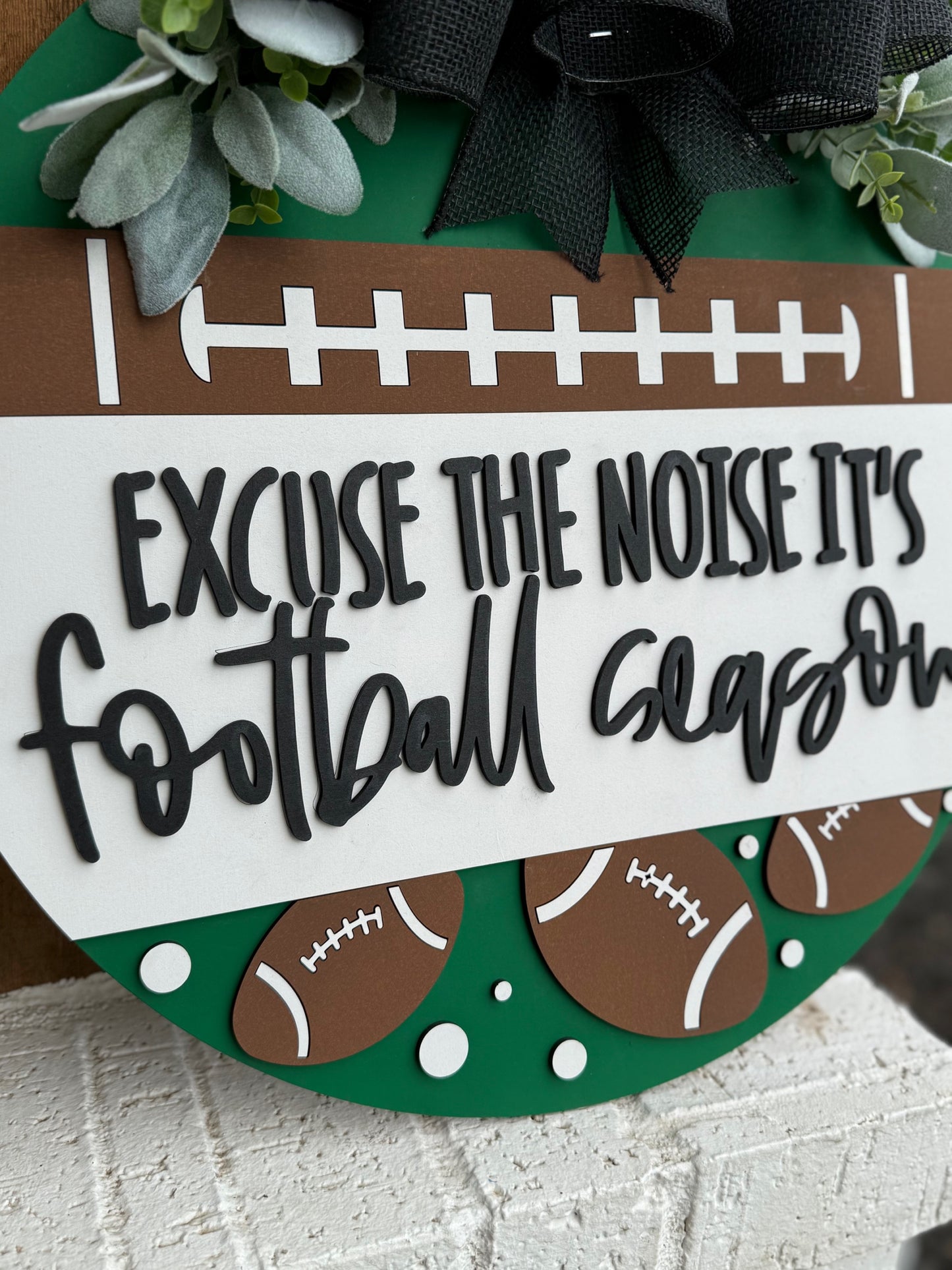 A round sign decorated with faux greenery and footballs reads, “Excuse the noise it’s football season” in bold, playful lettering. The sign features a football field design and is displayed against a white textured surface.