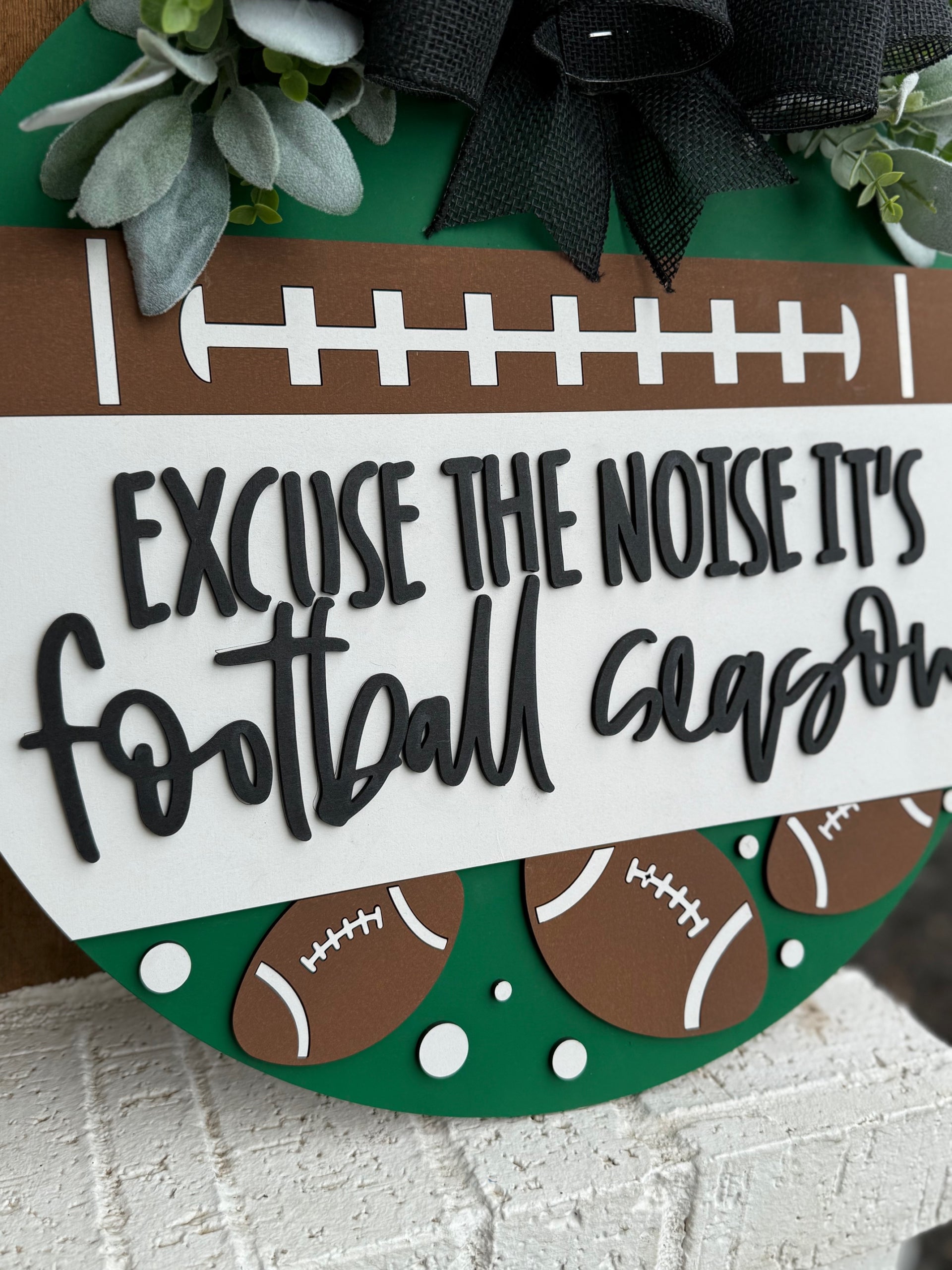 A round sign decorated with faux greenery and footballs reads, “Excuse the noise it’s football season” in bold, playful lettering. The sign features a football field design and is displayed against a white textured surface.