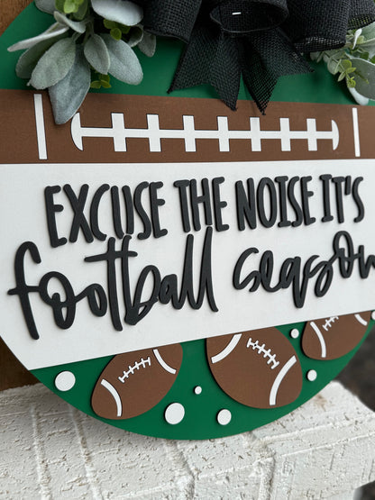 A round sign decorated with faux greenery and footballs reads, “Excuse the noise it’s football season” in bold, playful lettering. The sign features a football field design and is displayed against a white textured surface.