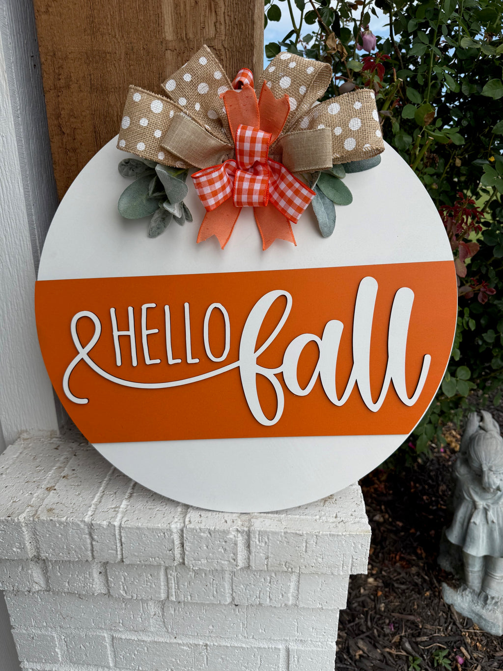 A round sign with Hello Fall in white script on an orange stripe, decorated with polka dot and checkered bows and faux greenery, is displayed on a white brick ledge near a garden.