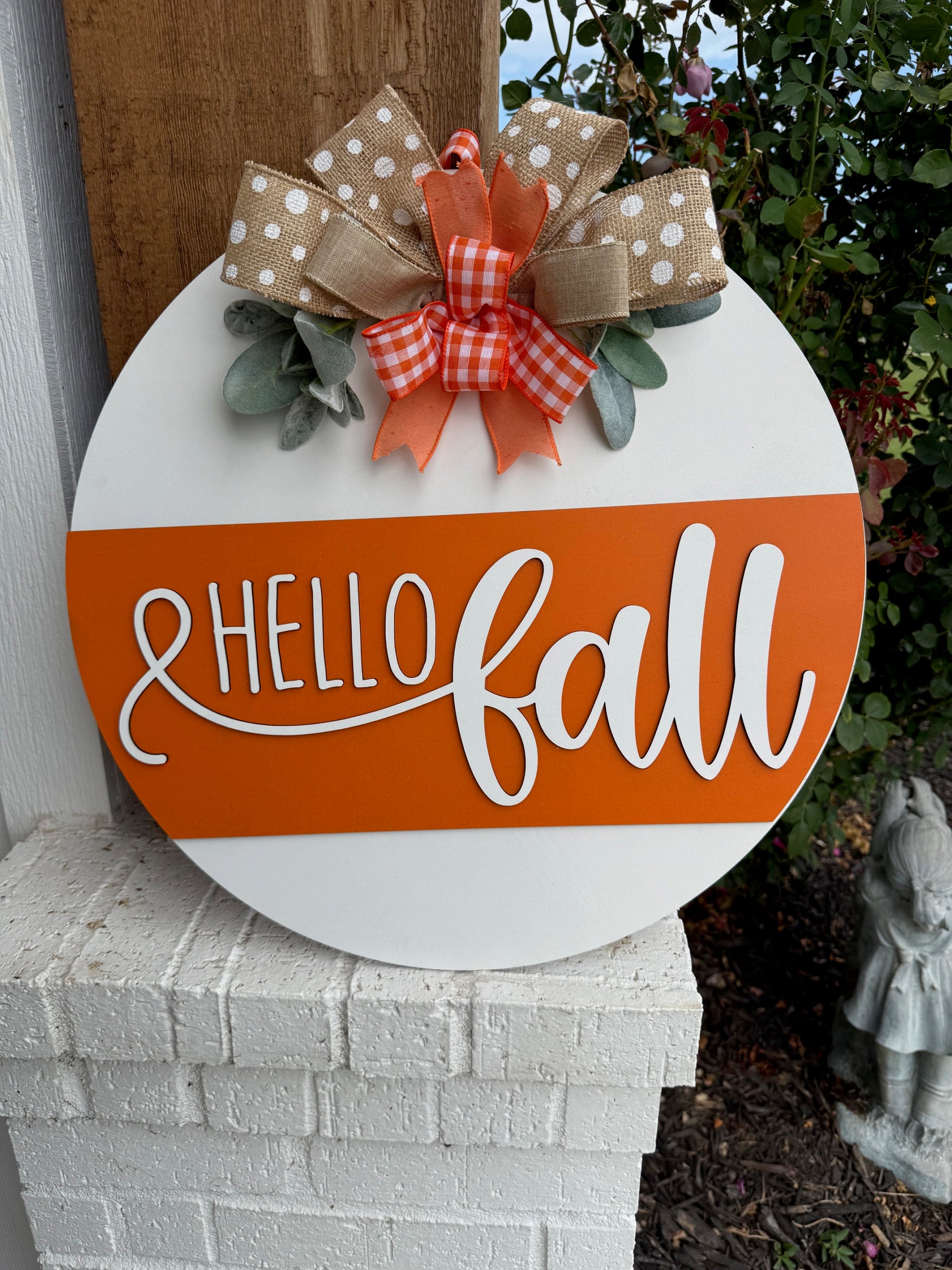A round sign with Hello Fall in white script on an orange stripe, decorated with polka dot and checkered bows and faux greenery, is displayed on a white brick ledge near a garden.