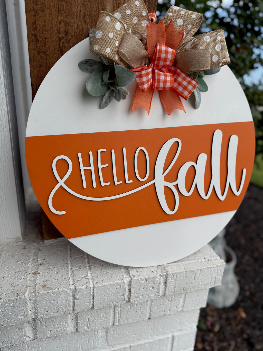 A round sign with Hello Fall in white script on an orange stripe, decorated with polka dot and checkered bows and faux greenery, is displayed on a white brick ledge near a garden.