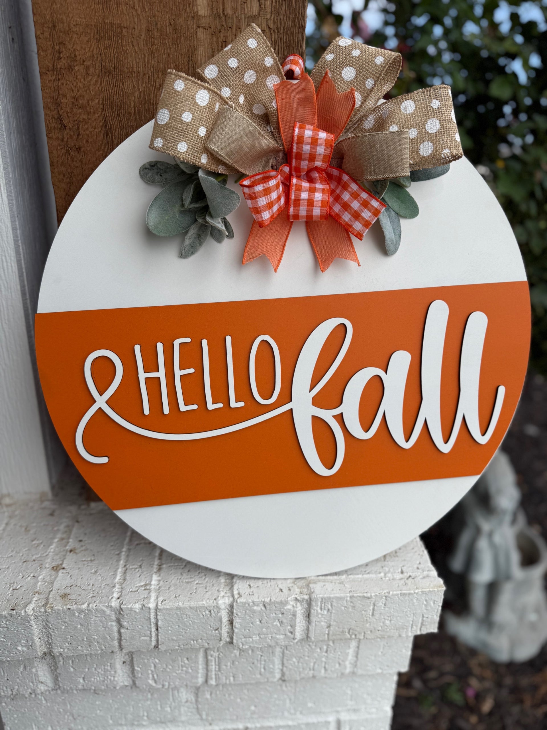 A round sign with Hello Fall in white script on an orange stripe, decorated with polka dot and checkered bows and faux greenery, is displayed on a white brick ledge near a garden.