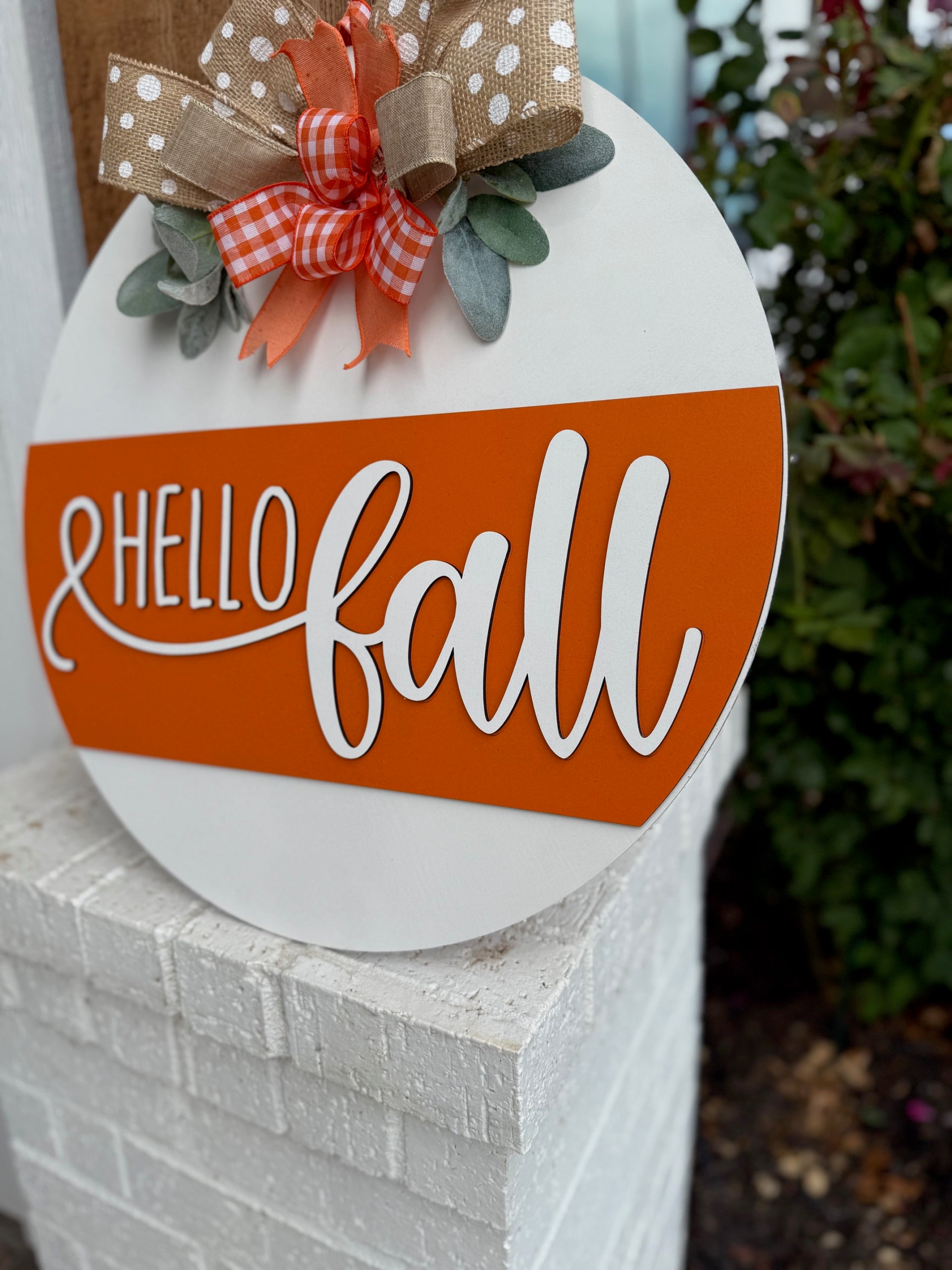 A round sign with Hello Fall in white script on an orange stripe, decorated with polka dot and checkered bows and faux greenery, is displayed on a white brick ledge near a garden.
