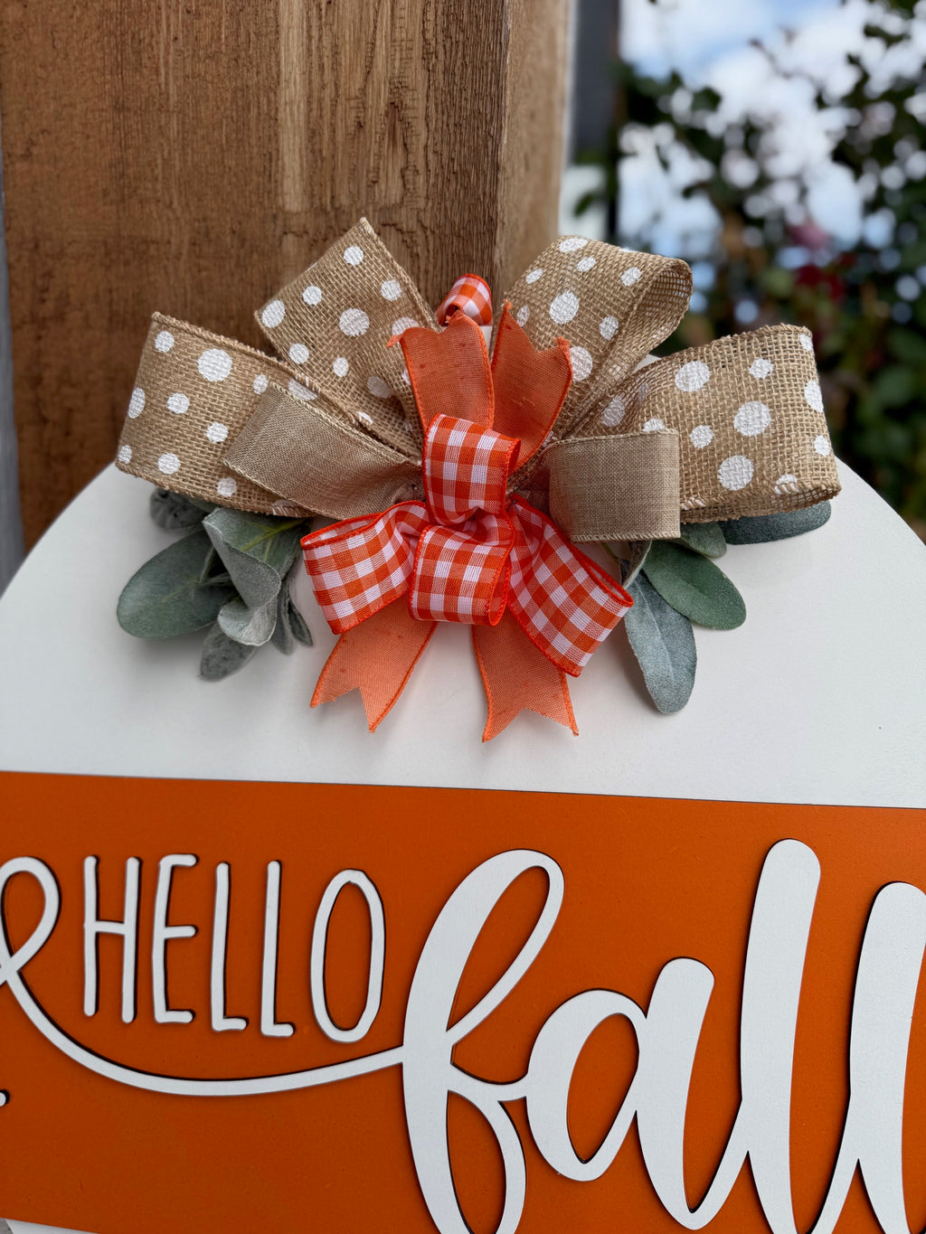 A round sign with Hello Fall in white script on an orange stripe, decorated with polka dot and checkered bows and faux greenery, is displayed on a white brick ledge near a garden.